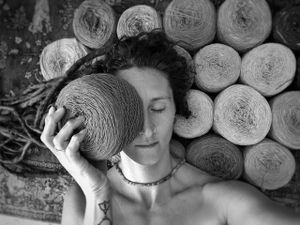 A black and white portrait of a woman laying with closed eyes on a rug with balls of yarn, holding a ball of yarn in fron.