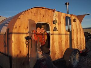 A man and woman sit in the door of a small wooden teardrop shaped trailer. 