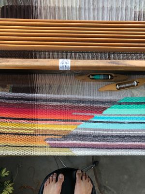 A view from above a loom, feet of the photographer on a black stool and a geometric rainbow of woven fabric on the loom