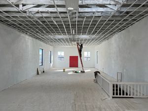 A woman is up on a red ladder in a huge white gallery space, white floor, ceiling and walls, with a red door at the back