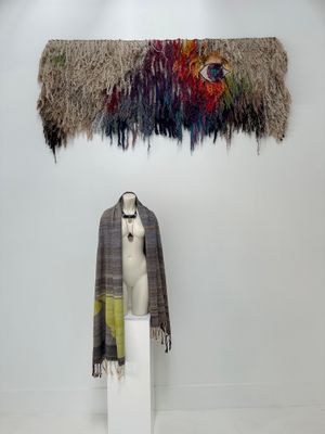 Two handwoven works of art, one rainbow and totally fringed, one subtle grey-blue and chartreuse on display in a gallery