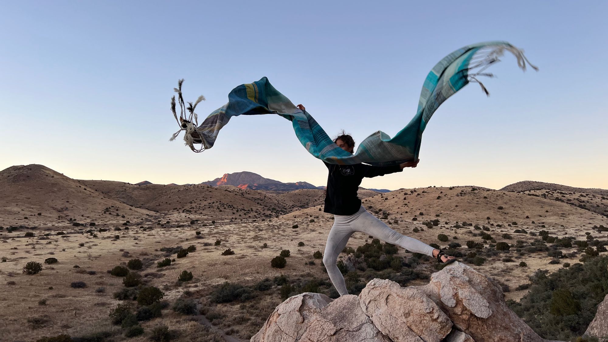 A woman stands on a rock out cropping holding a shawl which is floating on the wind. 