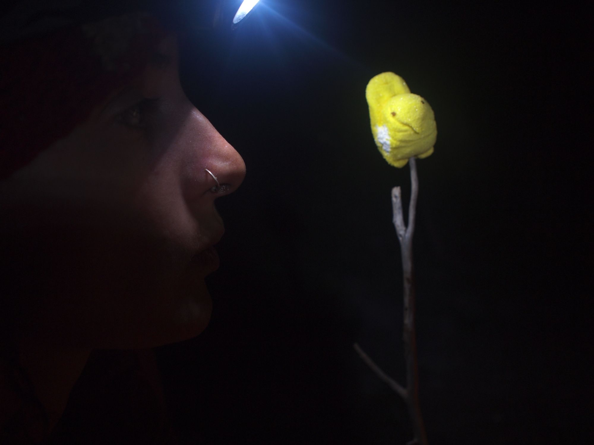 profile of a woman at night wearing a headlamp, holding a yellow marshmallow peep on a stick