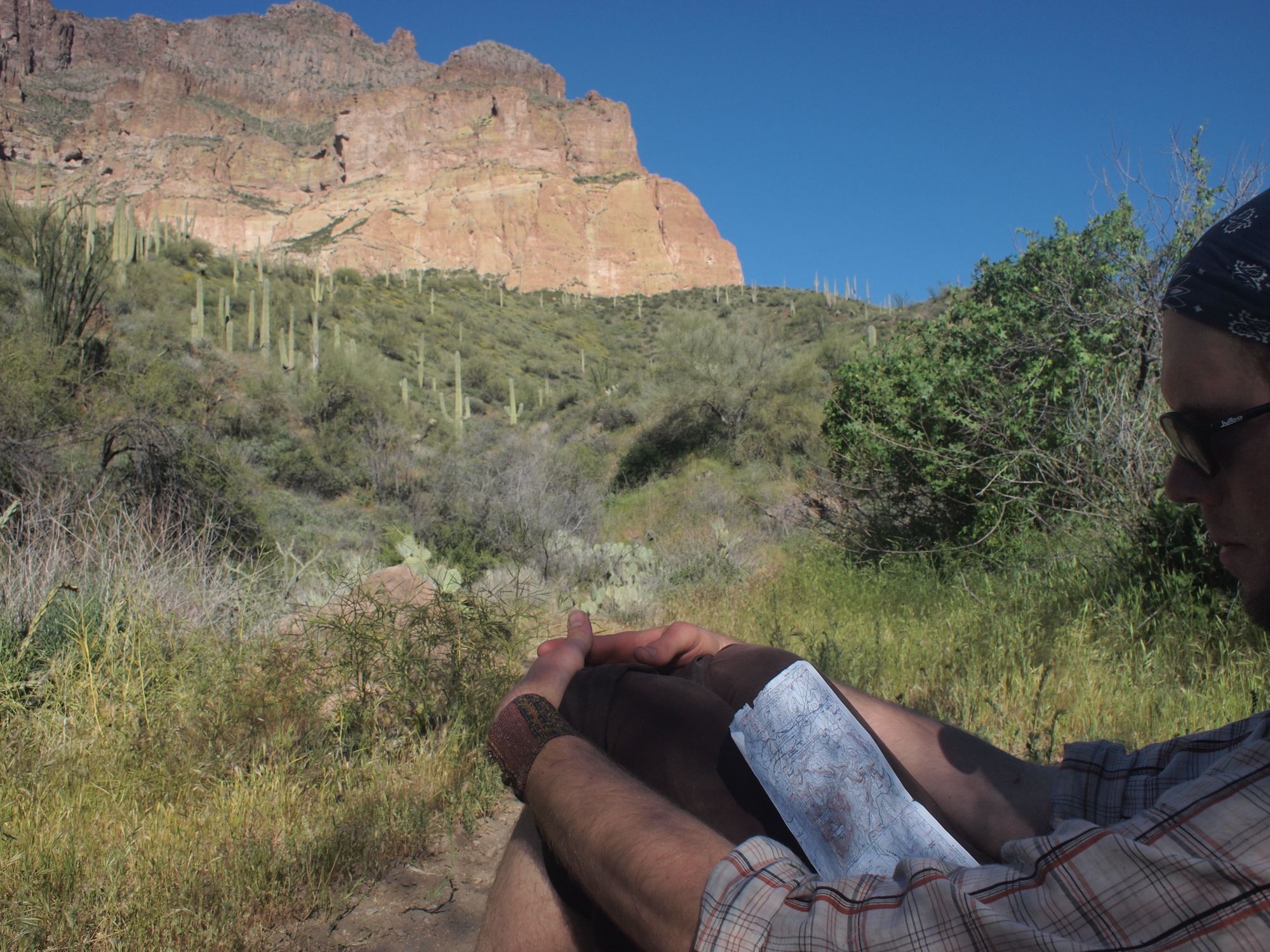 Man sitting by a trail looking at a topographical map with giant cliffs and cactus in the background 