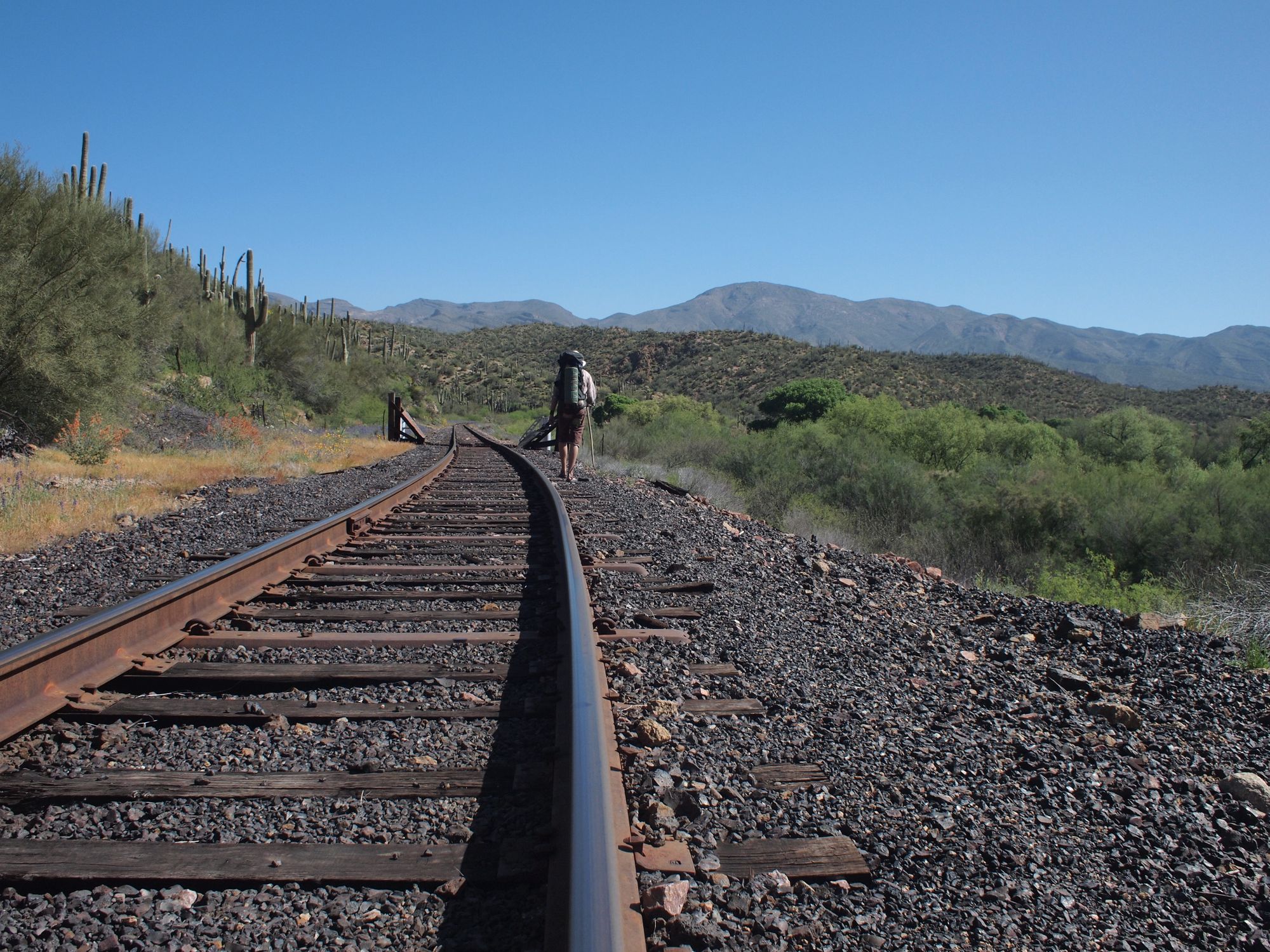 A man in a mountainous desert landscape, walking along railroad tracks. 