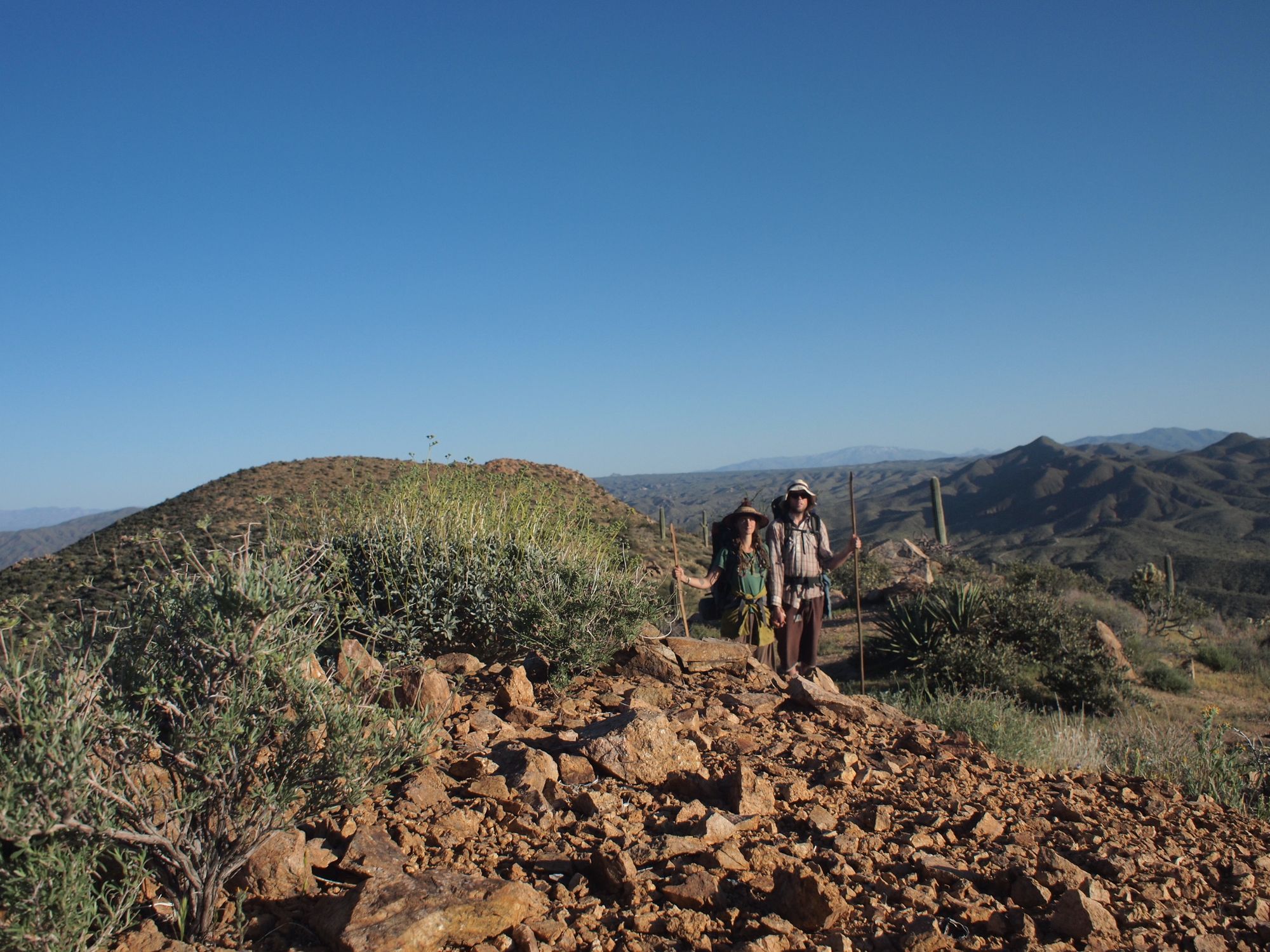 A man and woman stand, and in hand with walking sticks in a desert mountain landscape. 
