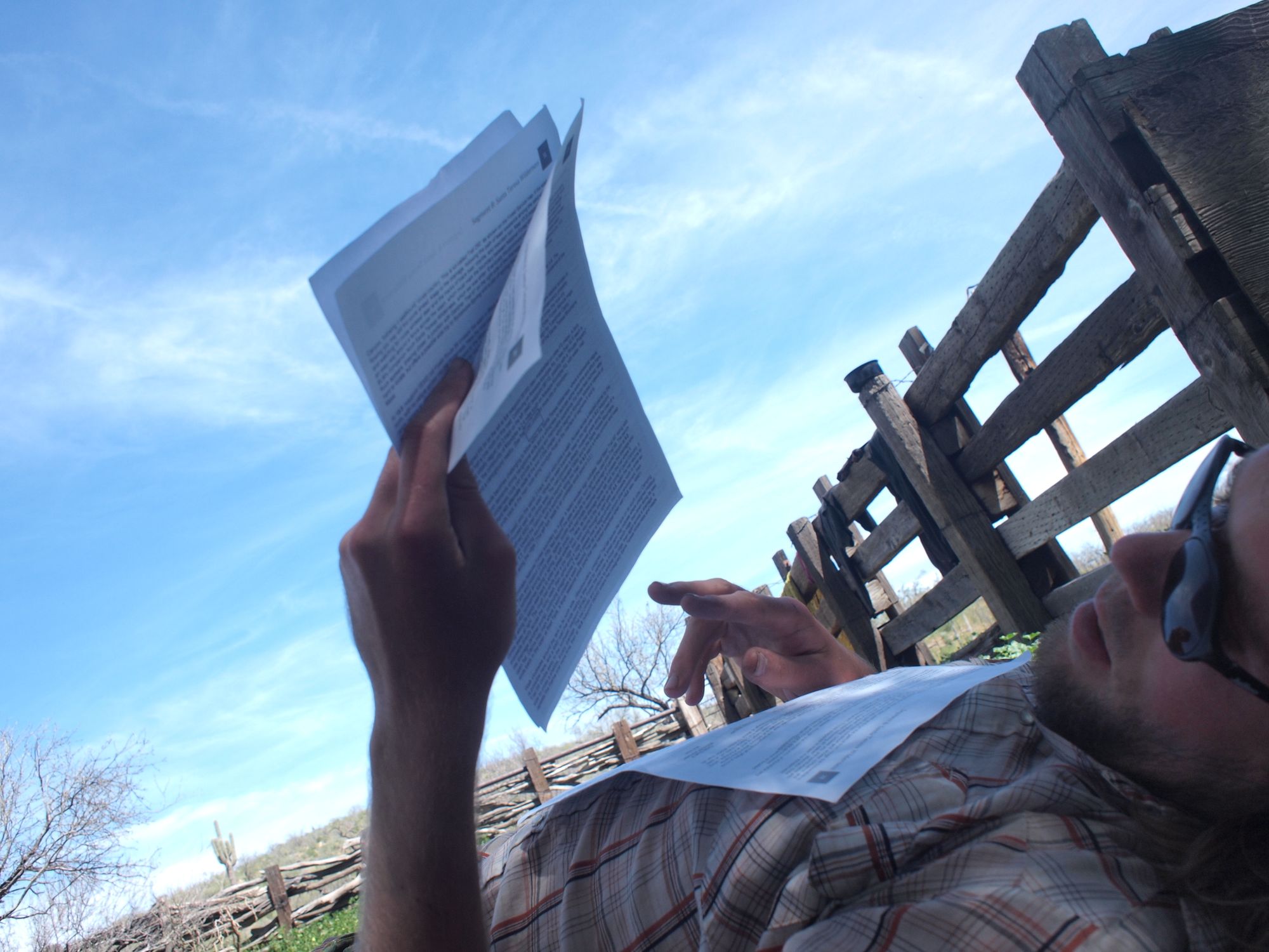 Man laying on his back next to a wooden fence, reading sheets of paper. 