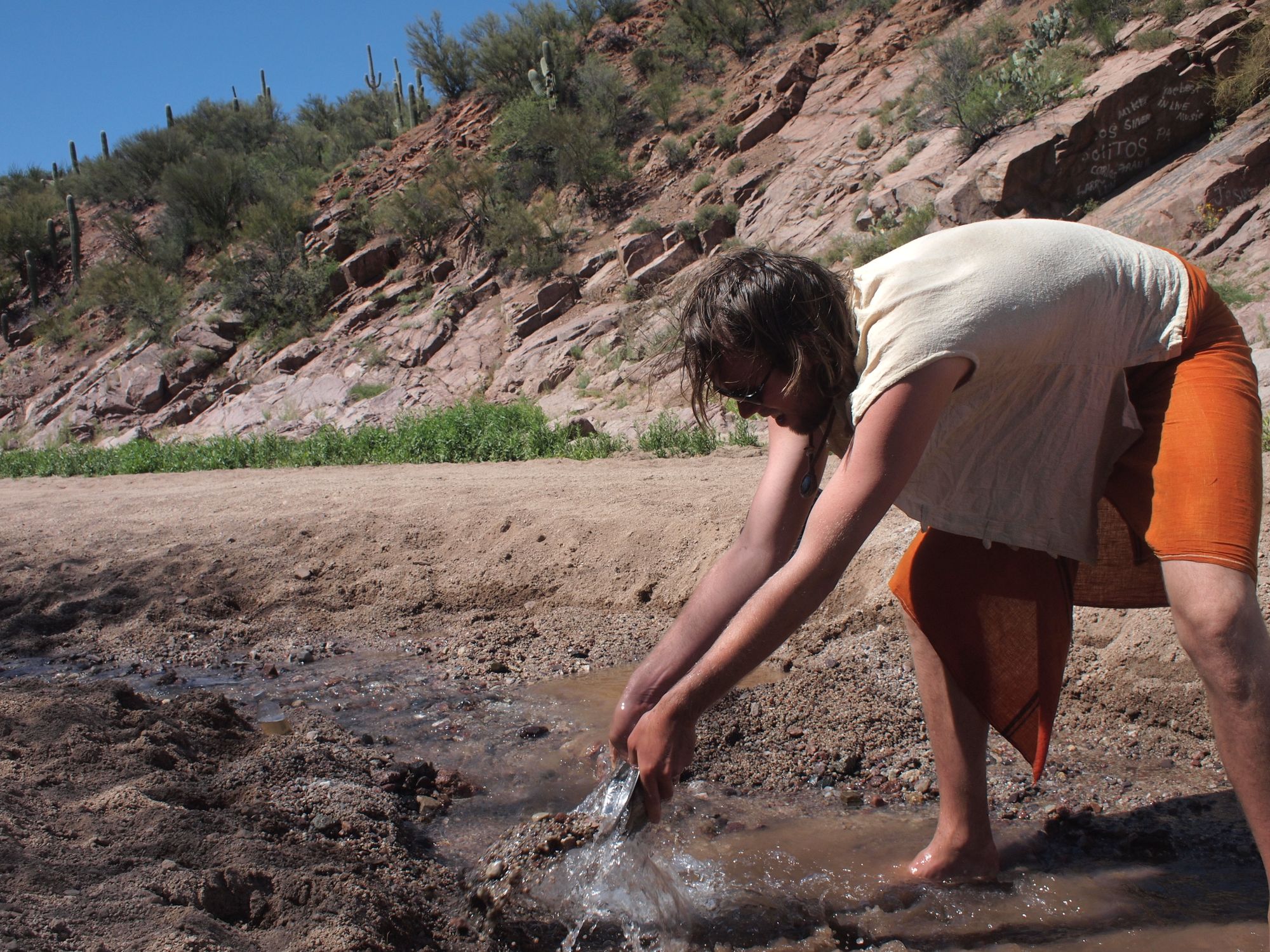 a man wearing a white shirt and orange sarong using a metal bowl to scoop sand out of a creekbed
