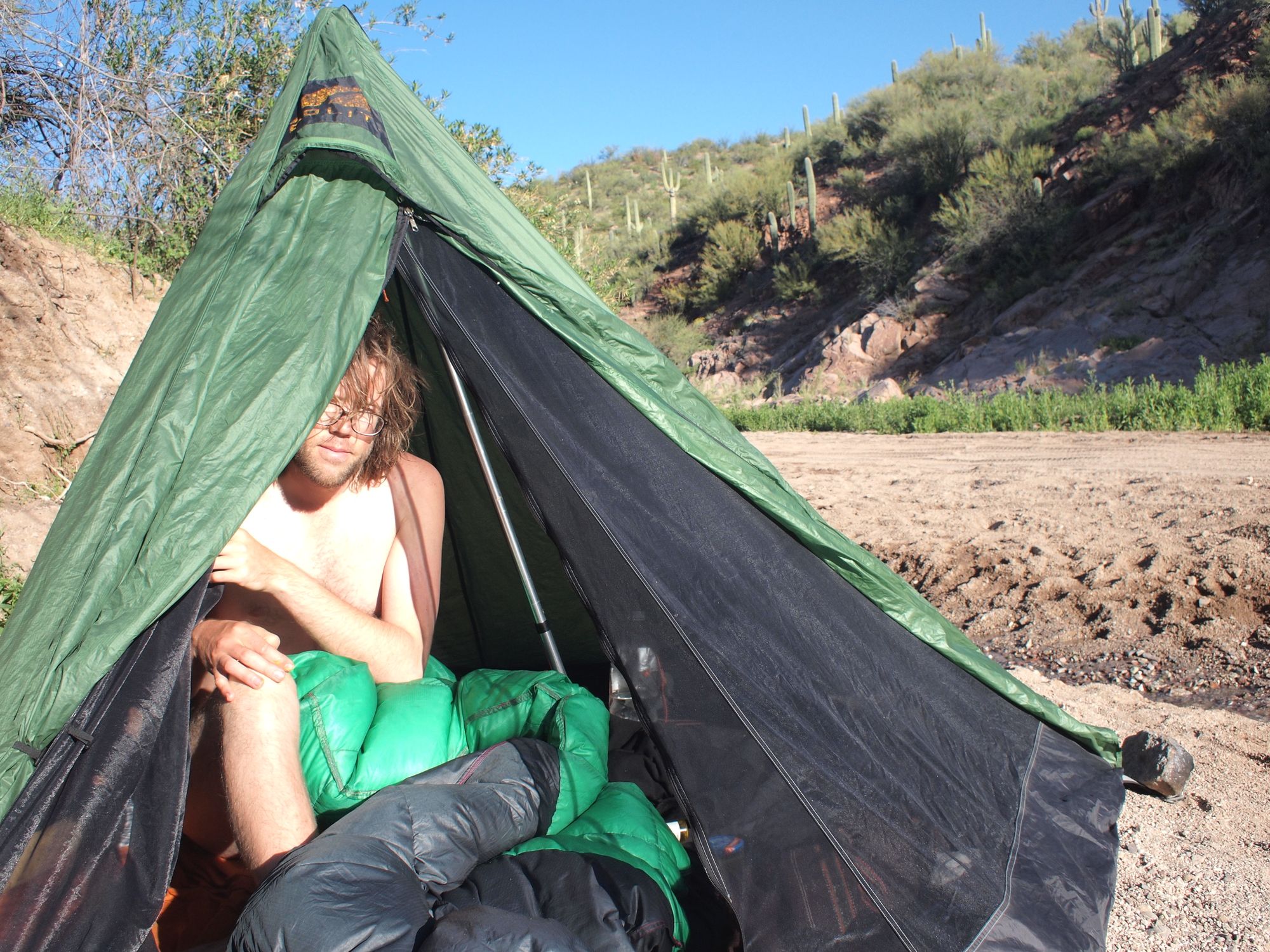 A man peeks out of a green and black tent in a sandy arroyo