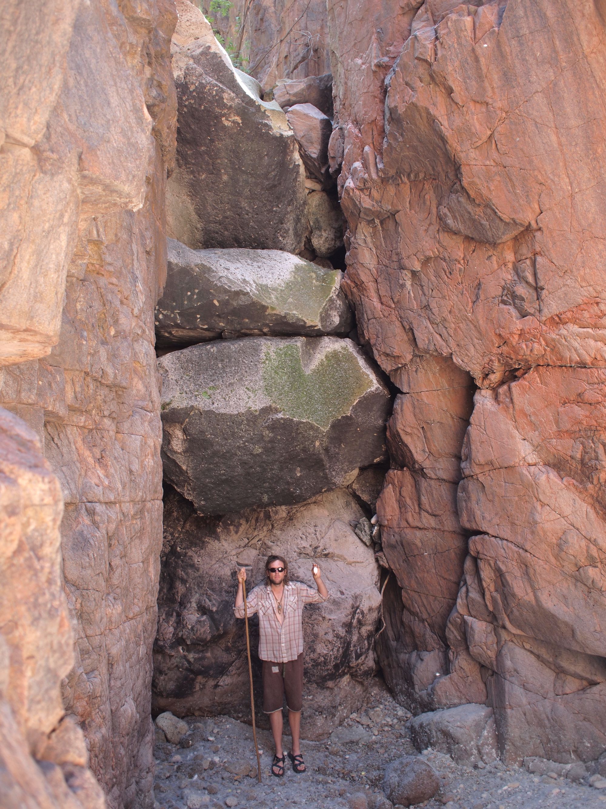 A man in plaid shirt and brown shorts stands in front of giant chockstone boulders blocking a canyon entrance 