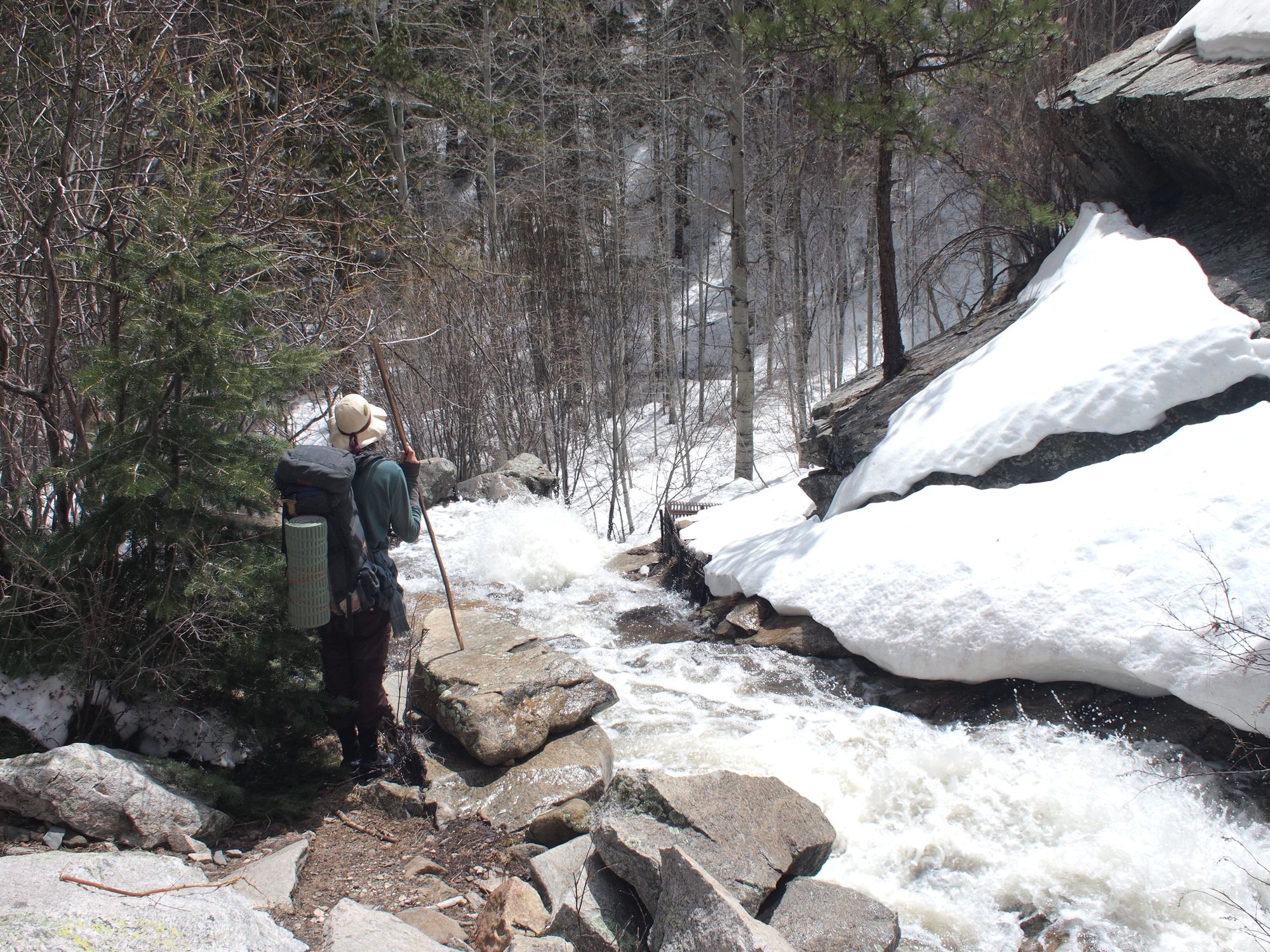 A man wearing a large backpack stands in a narrow canyon next to a raging whitewater creek liked with pine trees and snow