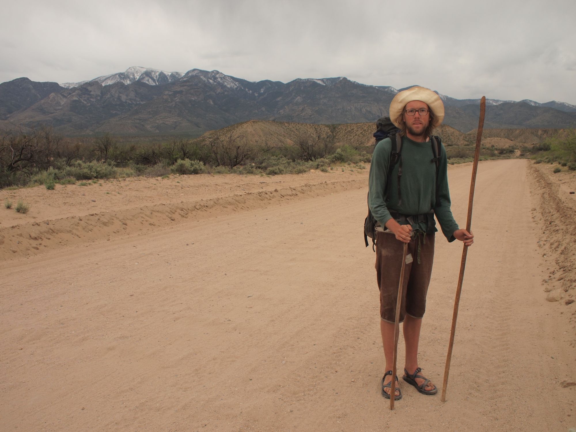 A man stands in the middle of a dirt road in the desert with snowy mountains behind. 