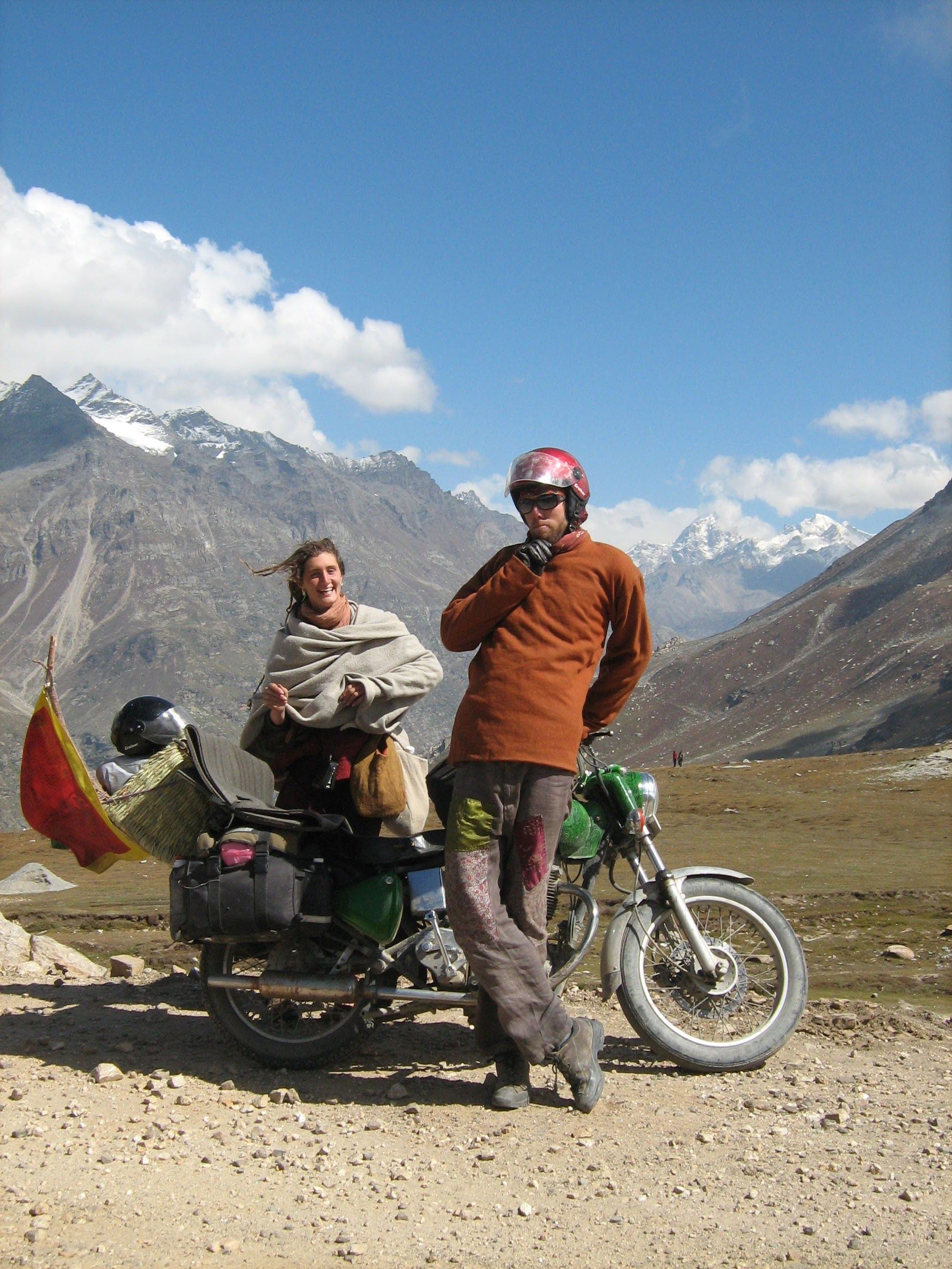 A man wearing an orange wool jacket and grey pants stands with a woman wearing a white blanket next to a green motorcycle, high on a mountain pass in the Himalayas. Snow capped peaks surround them. 