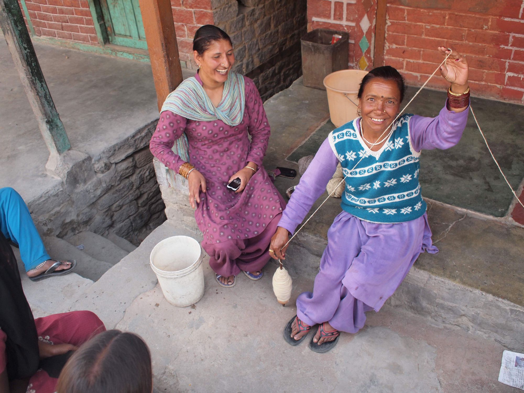 A woman in blue and white sweater vest and purple kurta spins wool and smiles at the camera, the woman next to her is wearing pink and teal and is laughing. 
