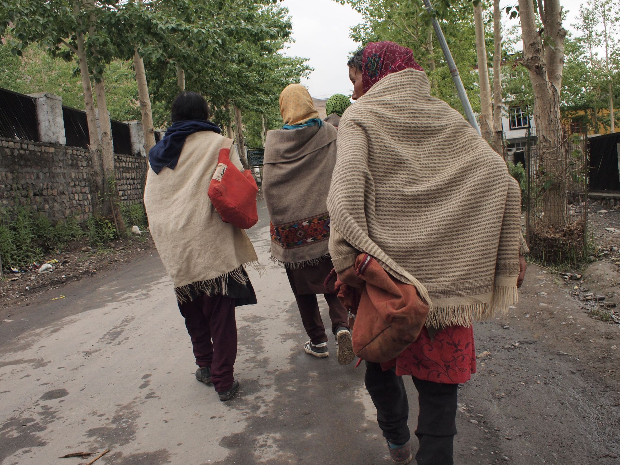 Three women in brown, tan, white blankets walk away from the camera and down a tree lined street. 