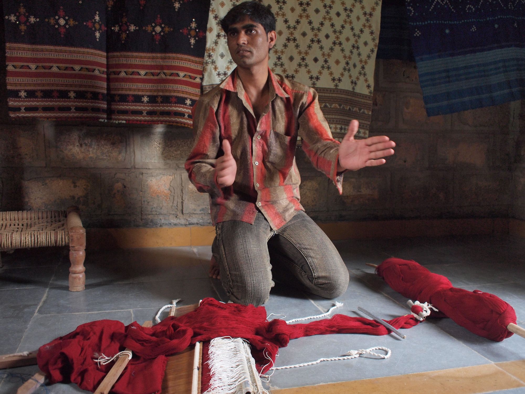 A man in red and brown shirt kneels on a grey stone floor, in front of him are pieces of a loom and a spool of bright red threads.