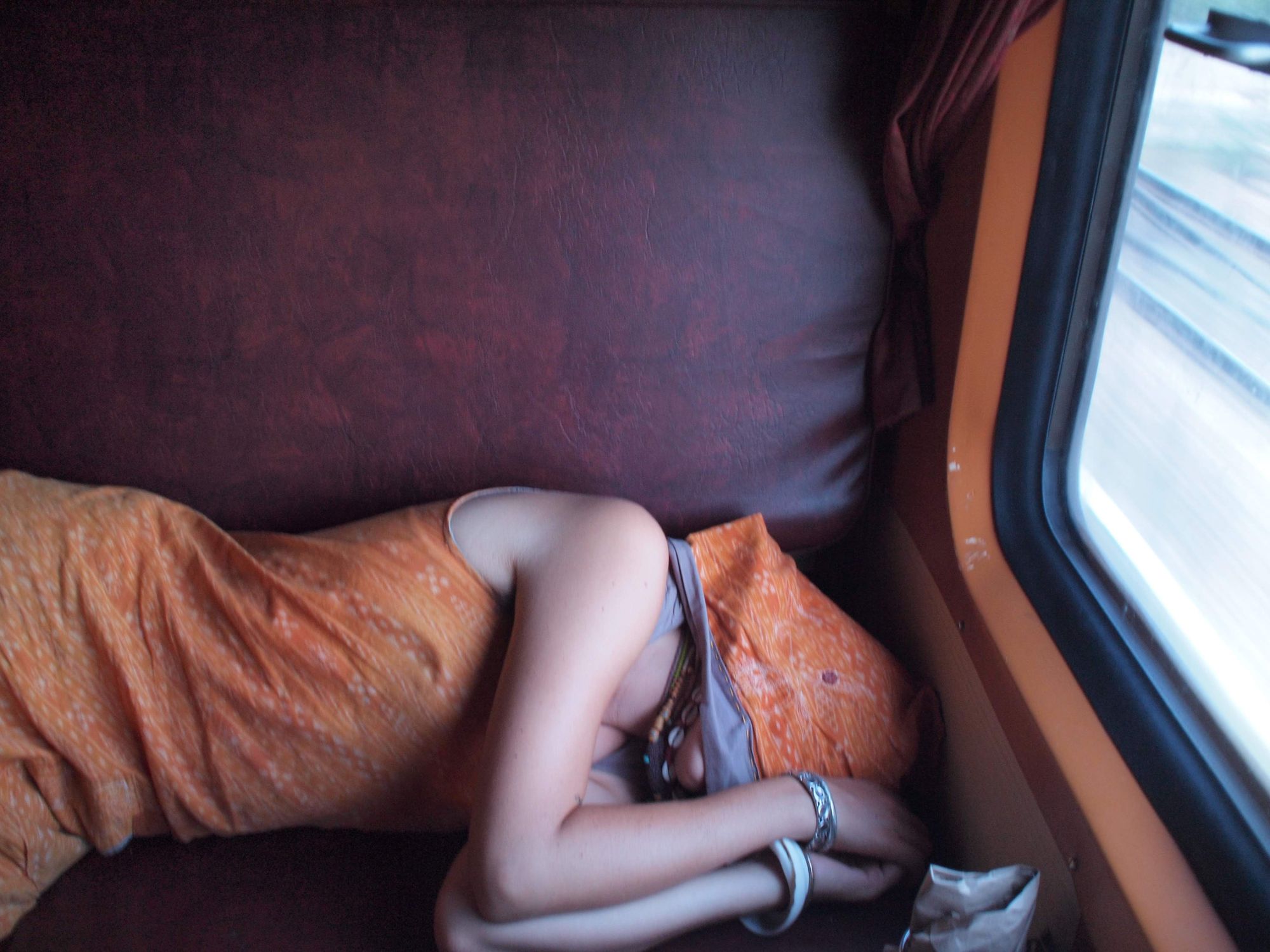 a woman in an orange dress with a large orange hood covering her face lays on a maroon vinyl train seat, napping. 