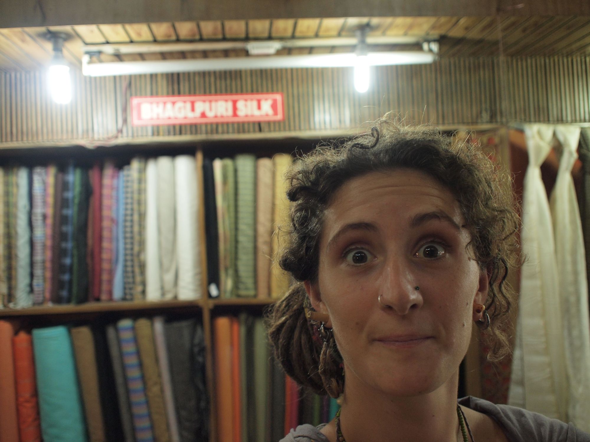 A woman with curly brown hair looks at the camera, making a funny excited face. behind her is a wall full of bolts of fabric of every color and a sign that says bhaglpuri silk