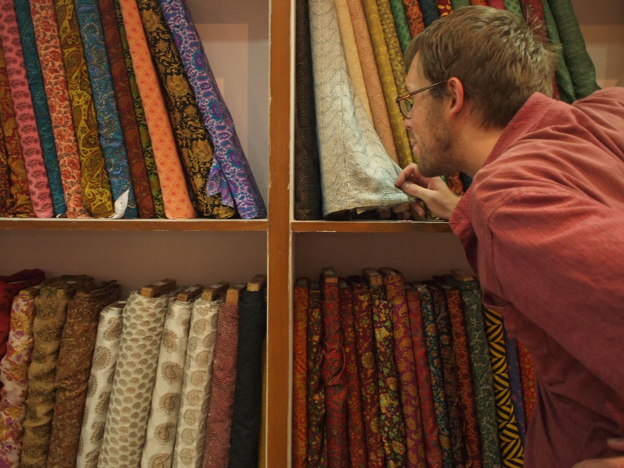 A man with glasses looks closely at white and blue fabric that is stacked on shelves of bolts of fabric of every color. 