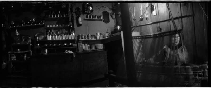 A black and white panoramic photo of a woman sitting at a loom in a kitchen, with many metal dishes on shelves. 