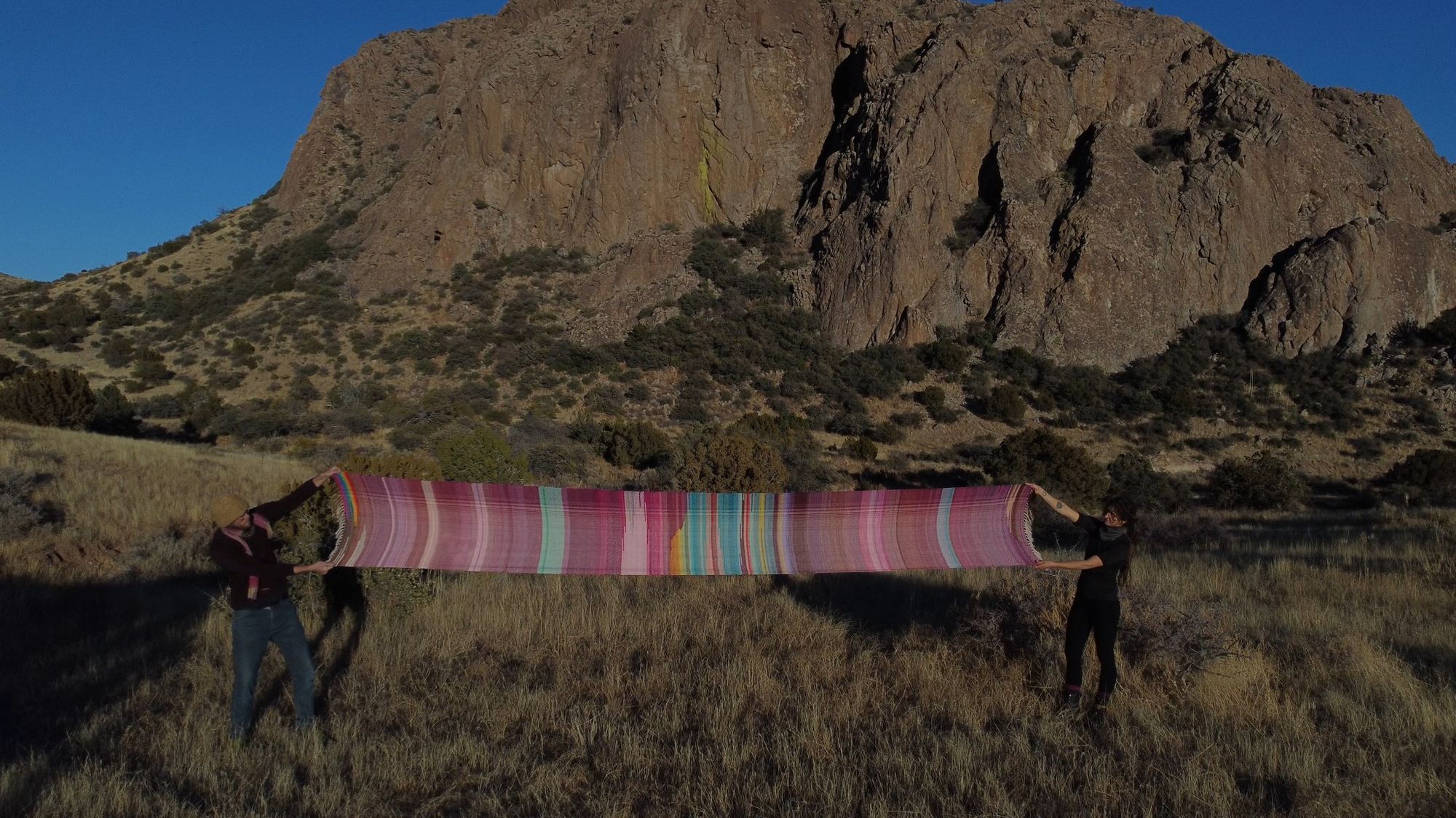 two people wearing black hold up a large piece of purple, orange, yellow, blue, rainbow and pink fabric in front of a large Rocky Mountain 