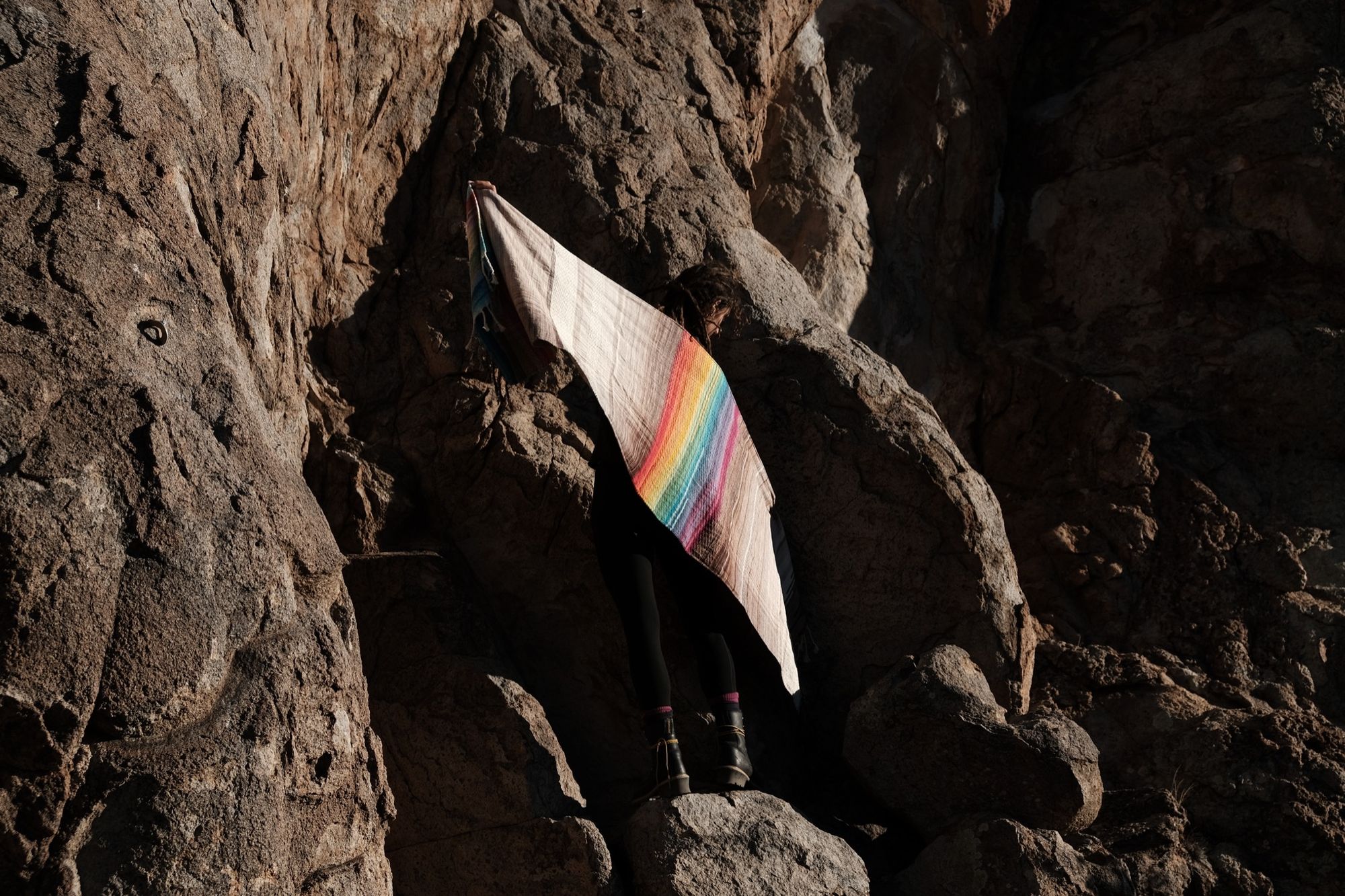A women stands in the shadow of a granite cliff silhouetted by evening light while holding a rainbow and grey handwoven shawl. 