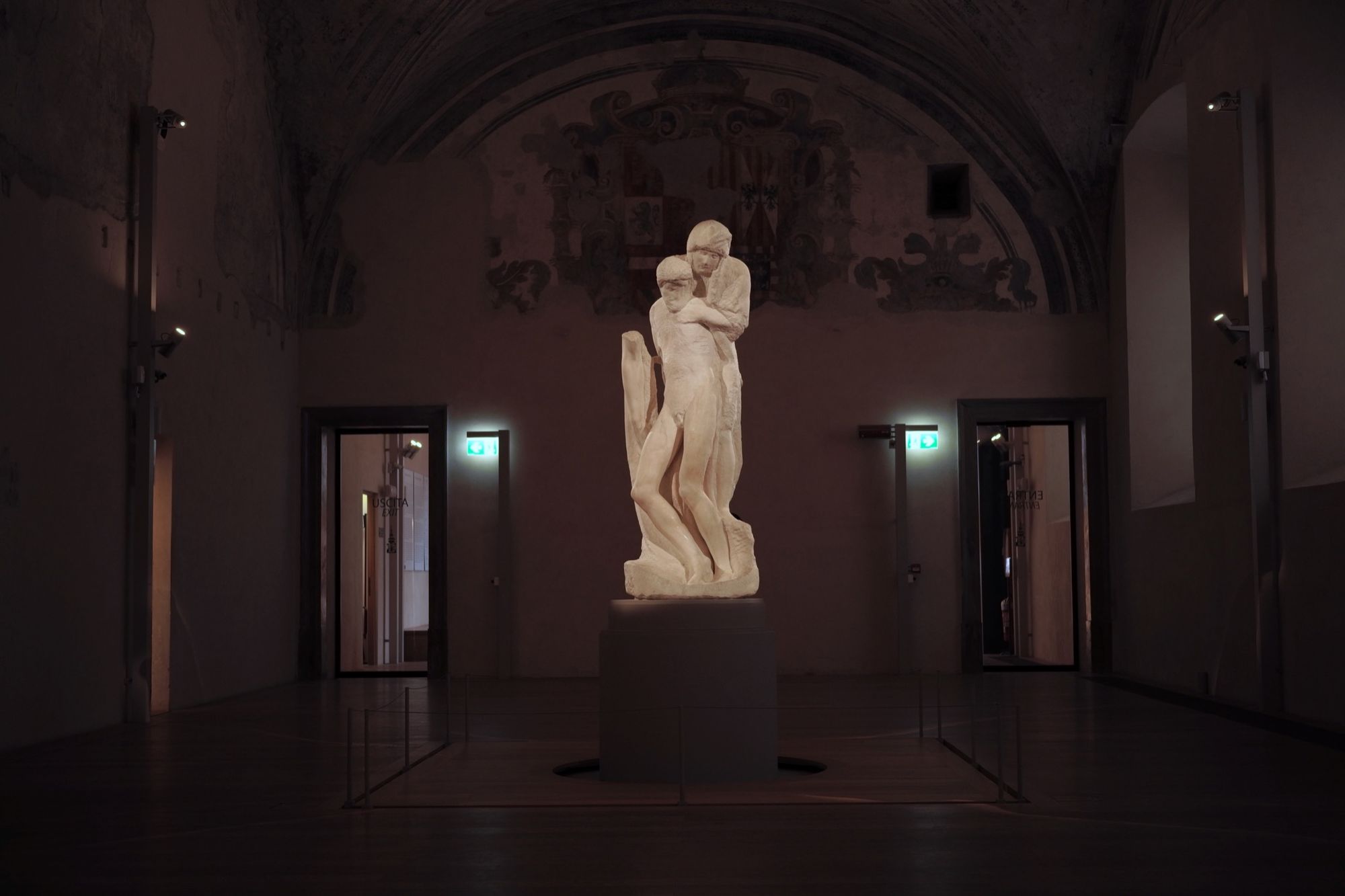 A sculpture of two figures, one holding the limp body of another, stands illuminated in center of a darkened room.