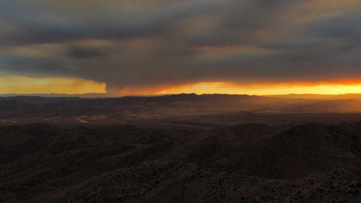 A landscape at sunset is draped in smoke with bright orange horizon and a massive column of smoke rises from a forest fire in the center of the image. 