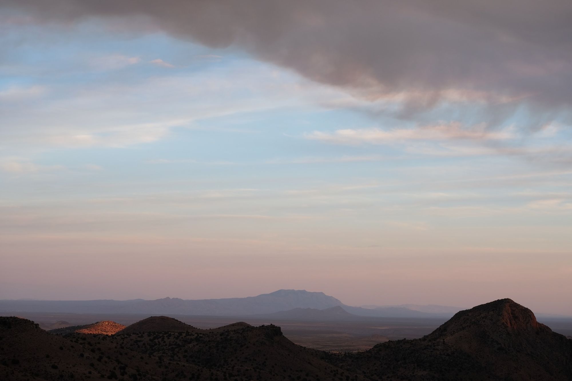 A panoramic view at sunset of the rio grande valley. Mountains in blue-grey shades recede into the distance while dark smoke pours in from above