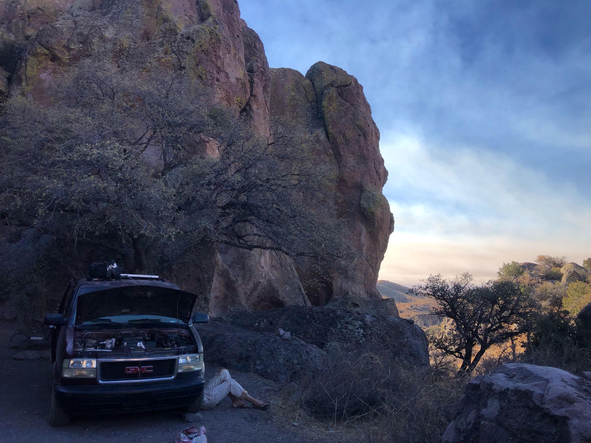 a maroon van has the hood up with a pair of legs popping out from underneath. It is parked under a scraggly oak tree with a canyon going down to the right. 