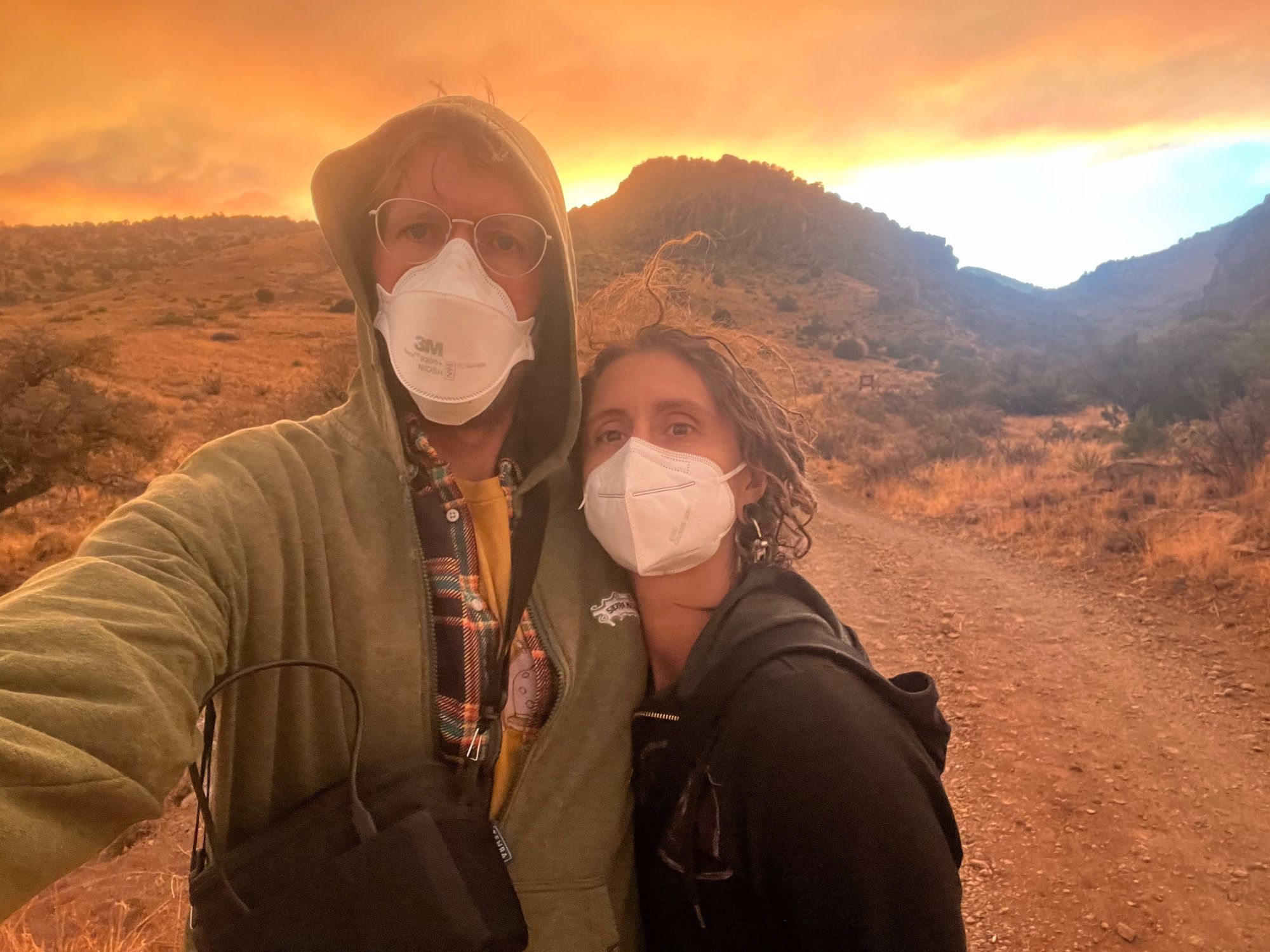 A man and woman stand on a dirt road wearing white facemarks, behind is a smoky red orange sunset
