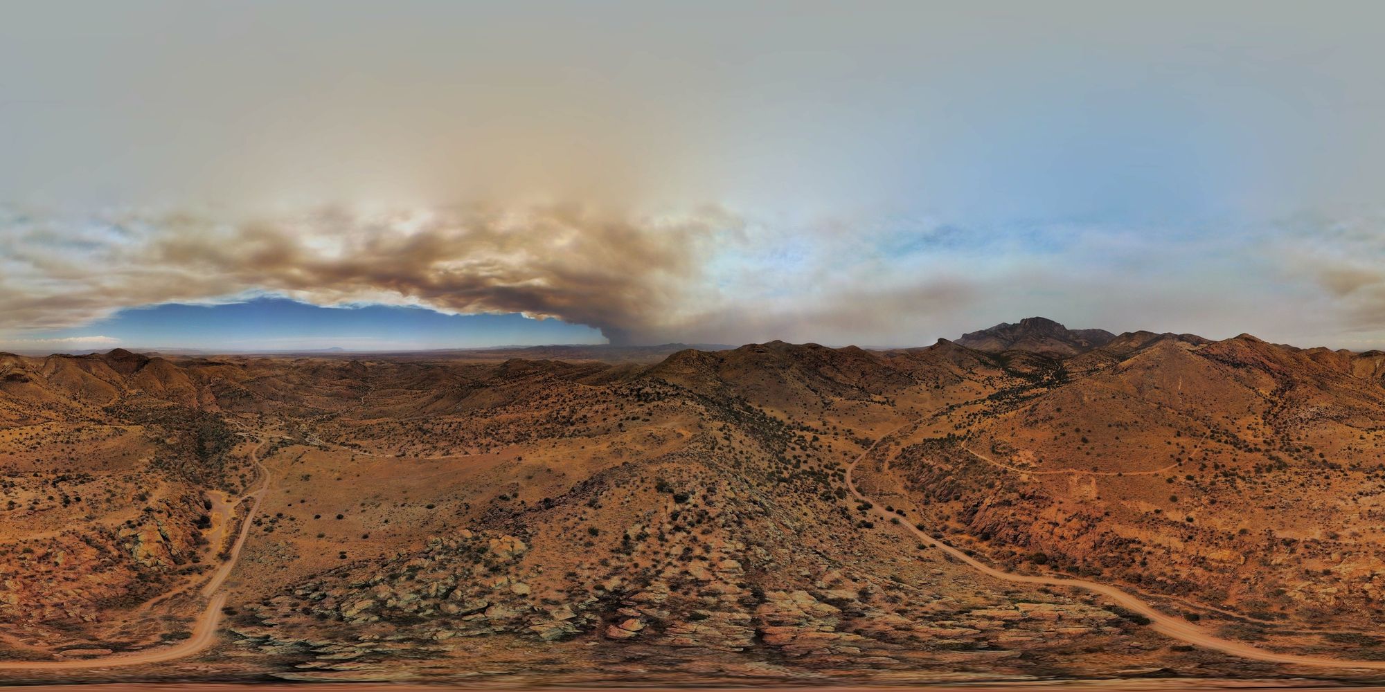 A desert landscape of hills and mountains with a giant column of smoke rising in the middle horizon with a plume of smoke filling the sky.