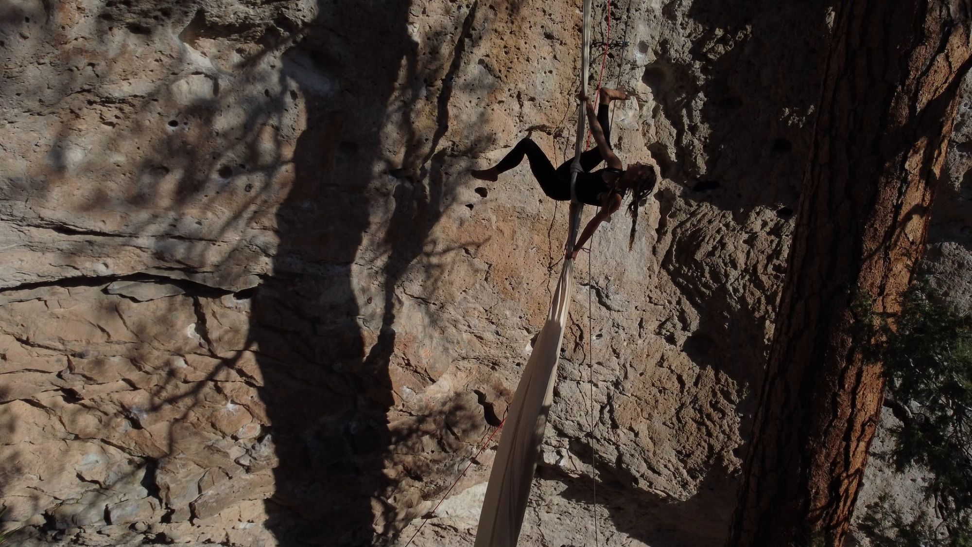 woman in a black unitard climbs white fabric hanging from a cliff, illuminated by bright sunset light. 