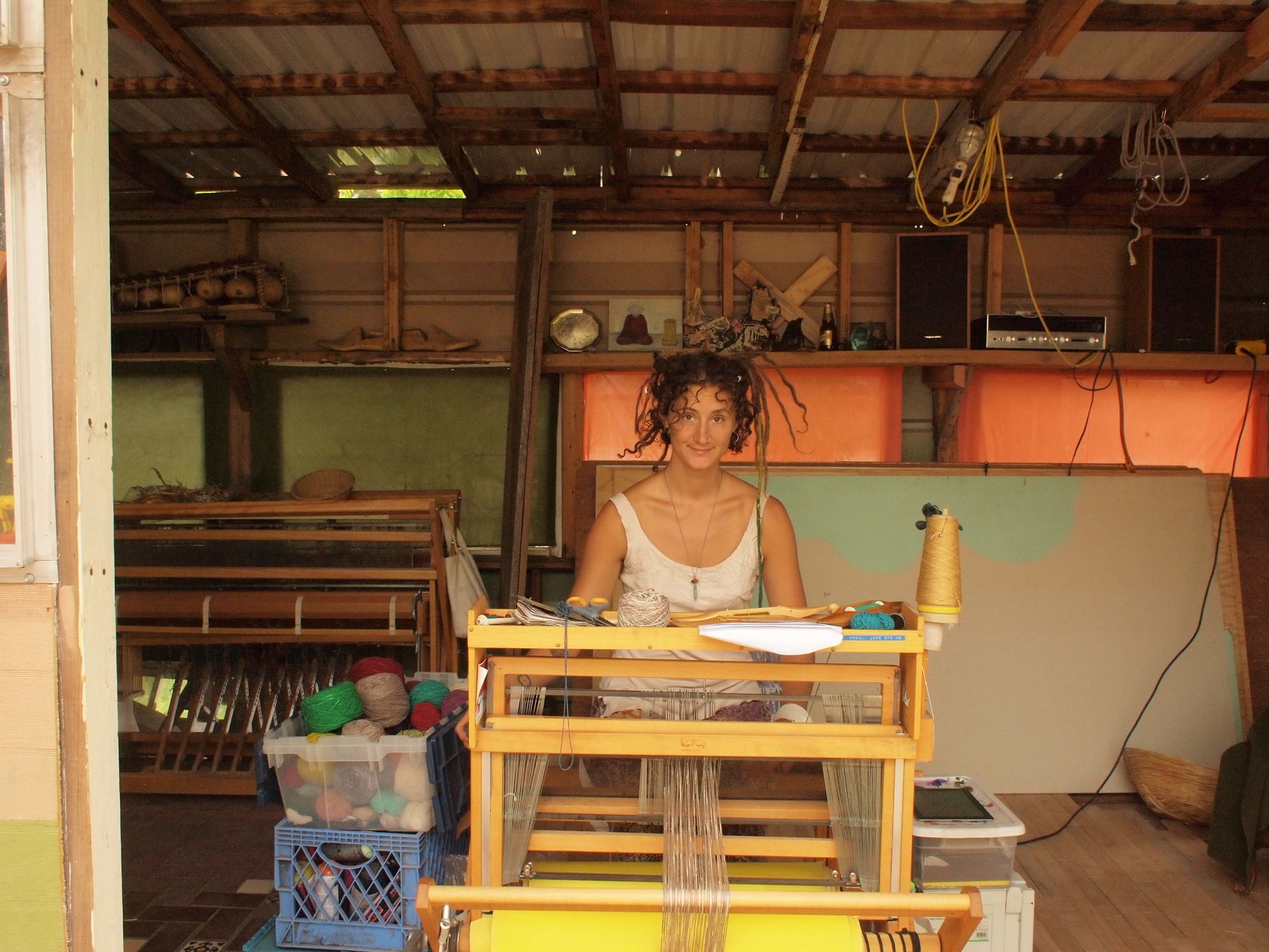 A woman sits at a loom weaving in an unfinished building with various objects on shelves behind her.