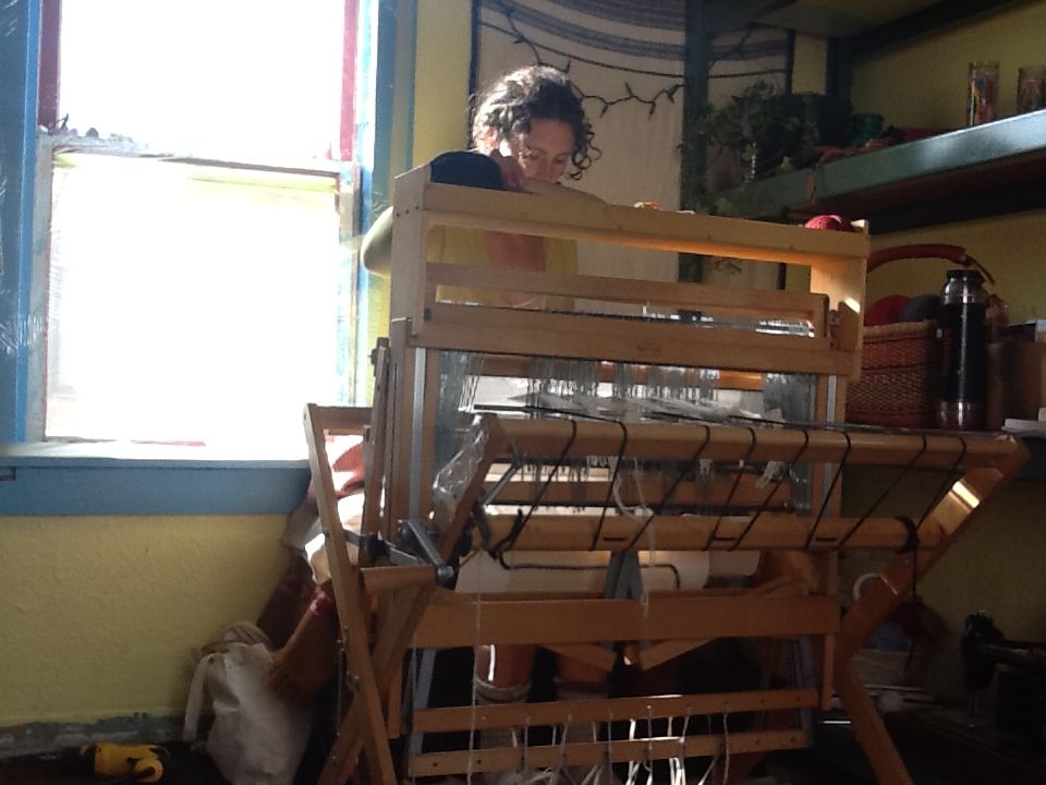 A woman sits at a loom weaving in the corner of a room next to a bright window. 