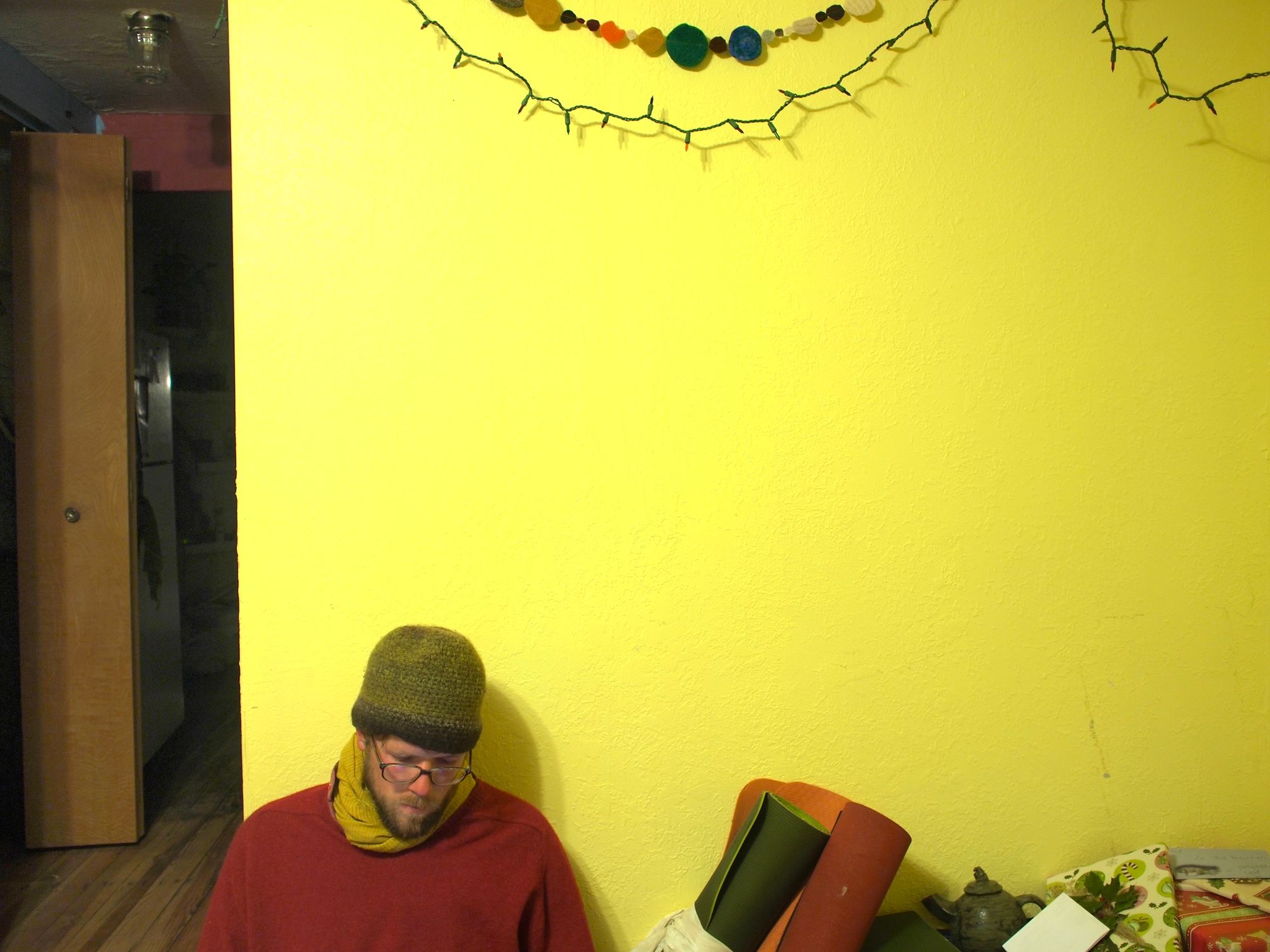 A yellow wall fills most of the picture while a man sitting on the floor looks down at his hands. 