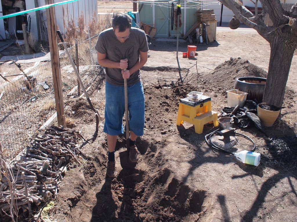 A man digs with a shovel in a dirt covered yard with a tree to one side and a shed in the background.