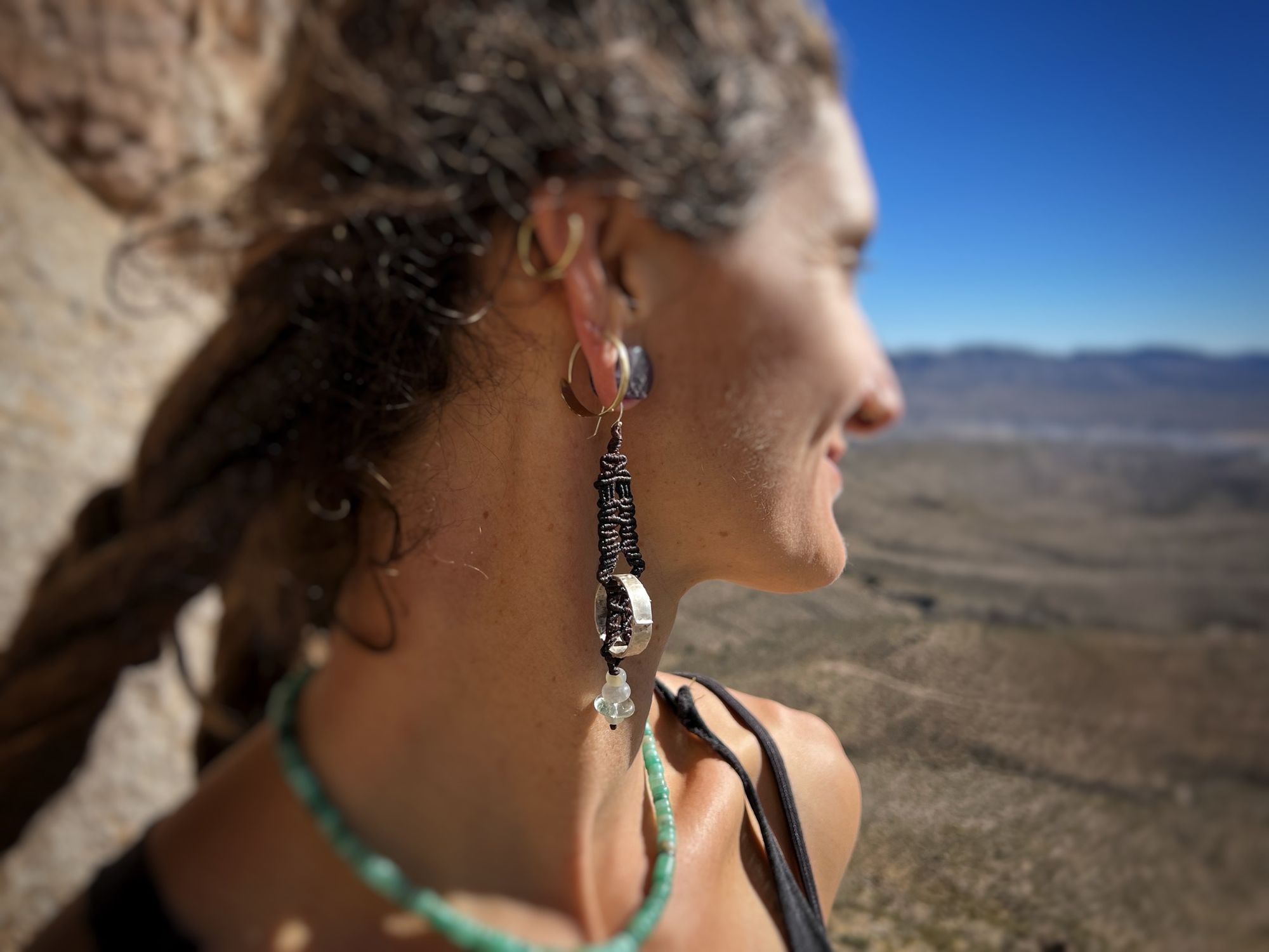 a woman in profile with a desert mountain landscape behind. she wears two gold hoop earrings and a purple knot work earring with a silver ring