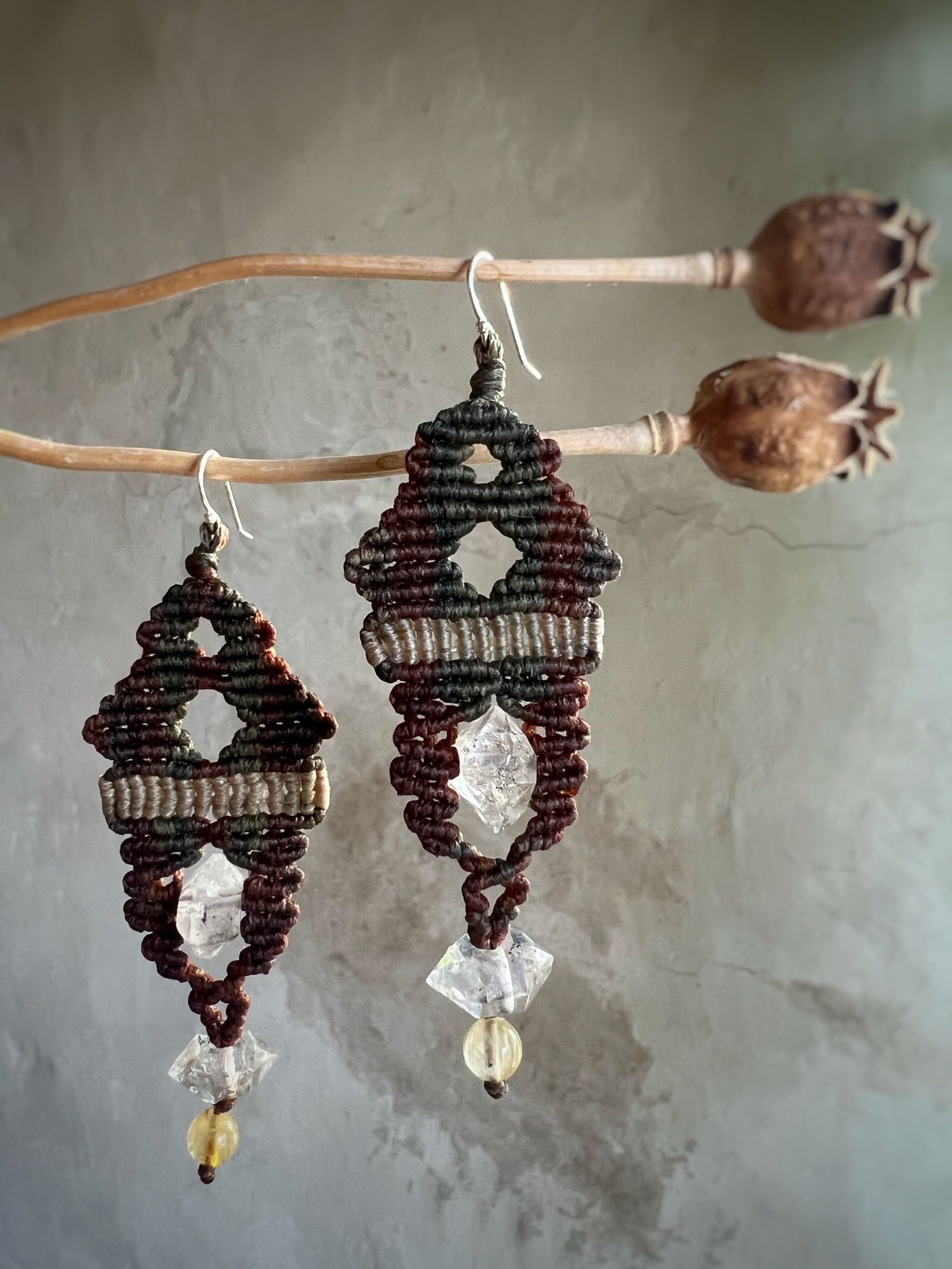 a pair of brown and green knot work earrings with clear double terminated crystals hang from dry poppy stems. 