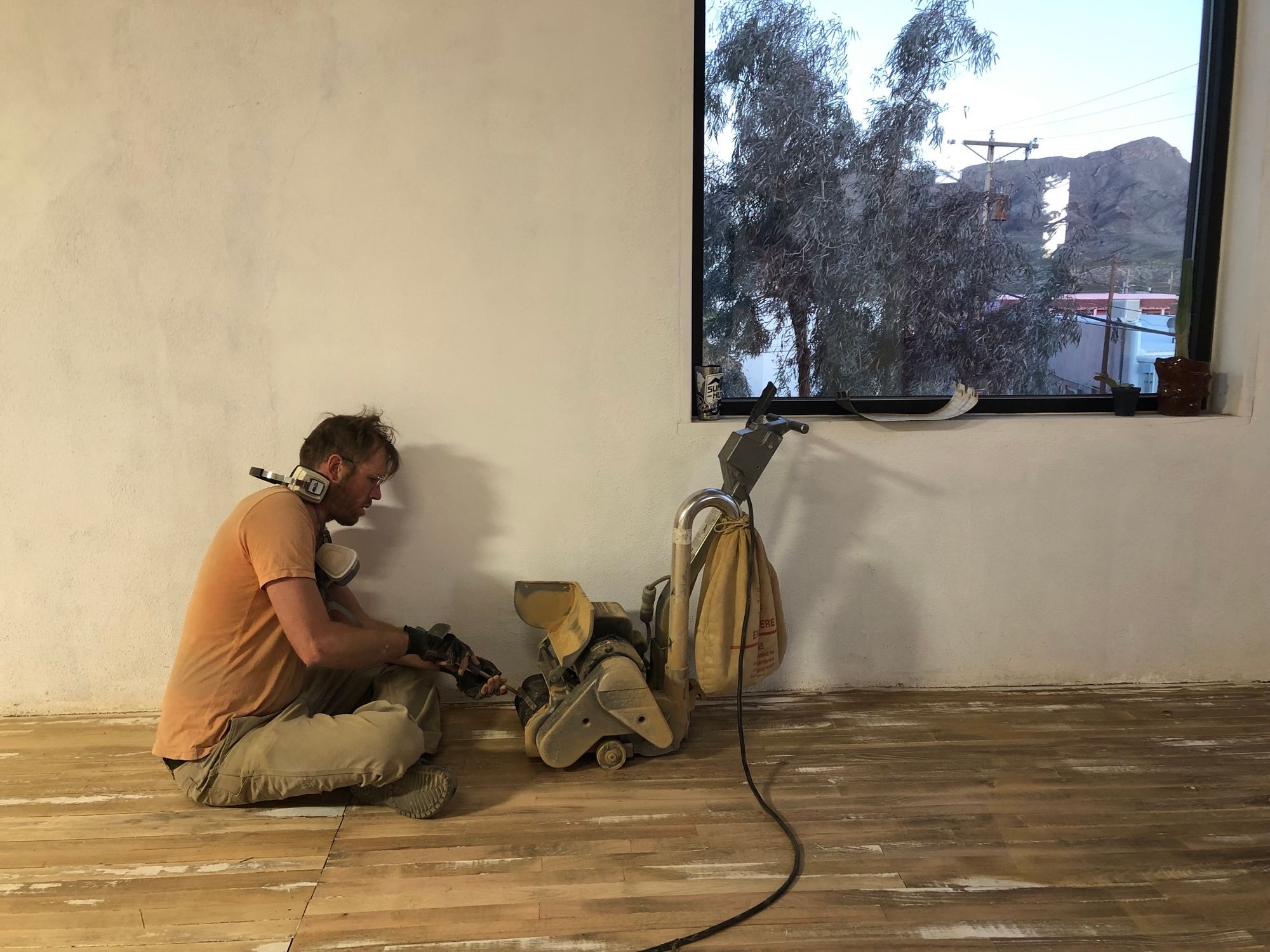 A man in an orange t-shirt and tan pants sits on a wooden floor on front of a white wall with a big window with a mountain view. He is changing the belt on a large drum sander