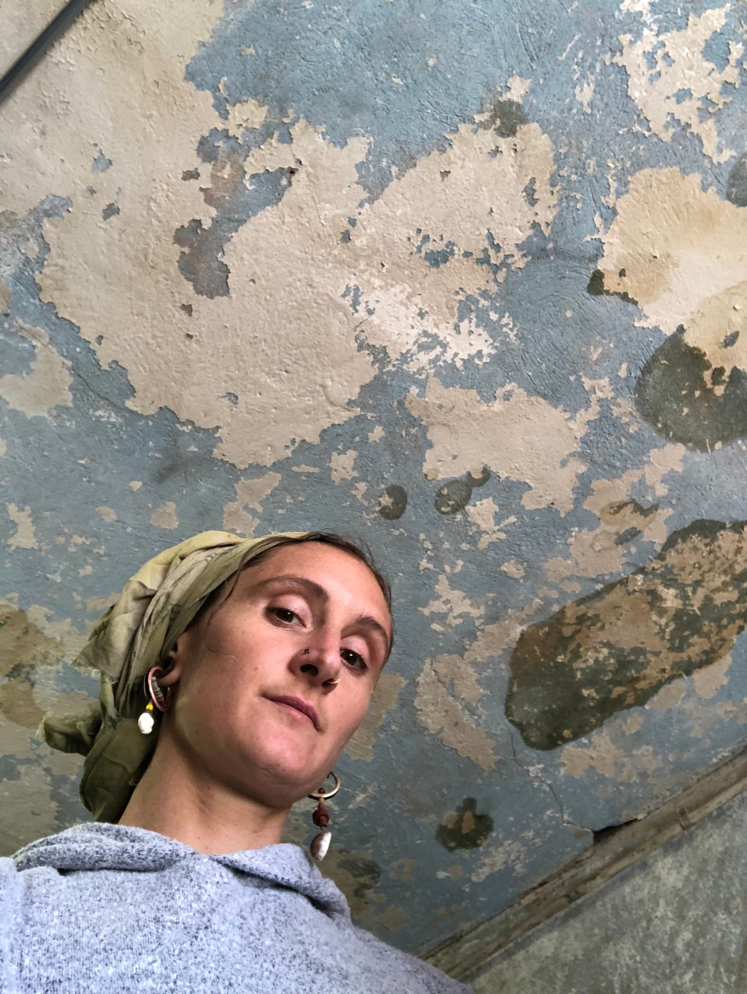 a view from below as a woman looks down at the camera wearing a green headscarf. The ceiling above her is white and blue peeling paint with water stains. 