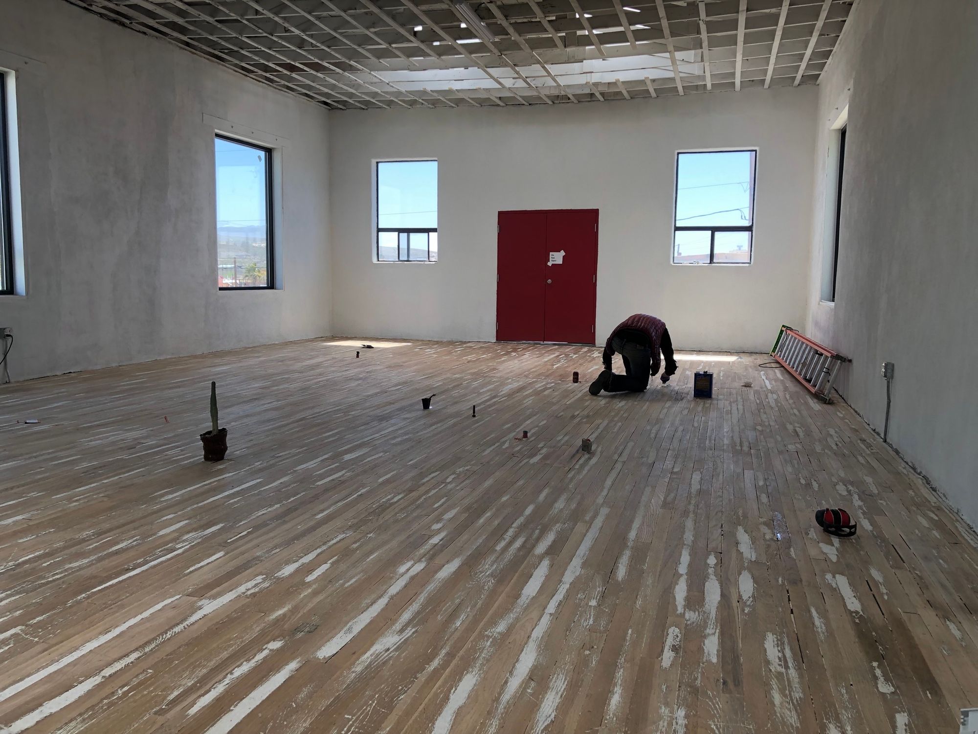  a man kneels on a wooden floor in a white room with big windows and a red double door.