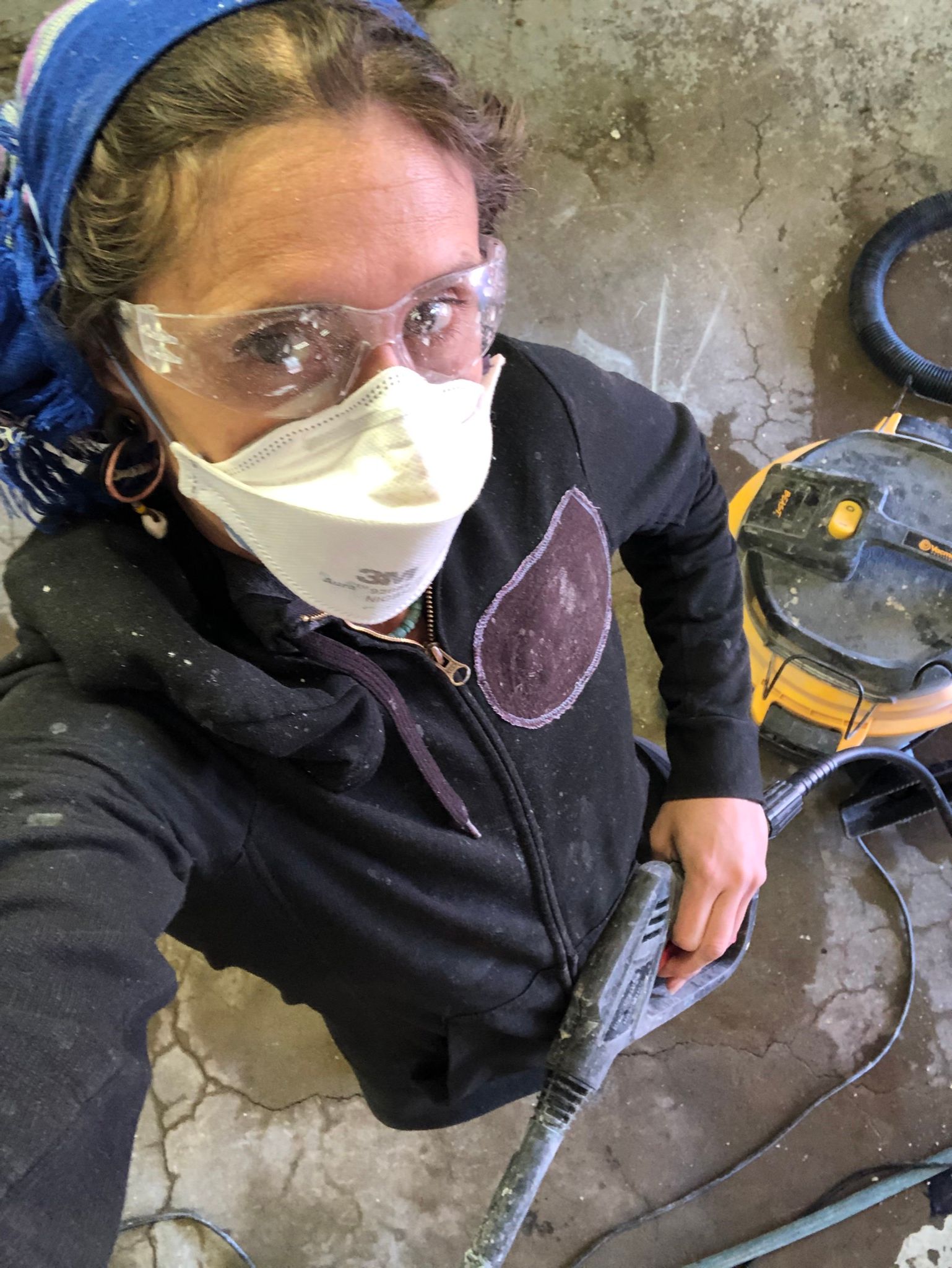 a view from above of a woman wearing ppe of a facemark, goggles and a blue headscarf. she holds a power washer in one hand and is covered in white paint.