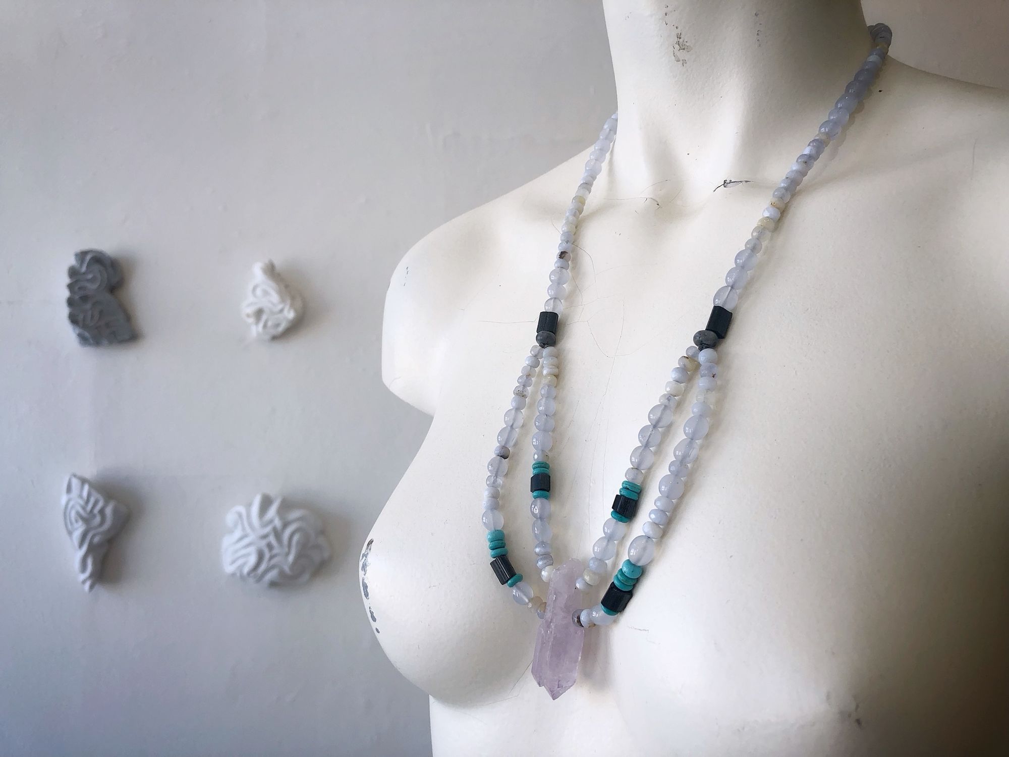 A white mannequin wears a necklace of pale blue, purple, turquoise and black semi precious stones in a gallery with marble sculptures on the wall 
