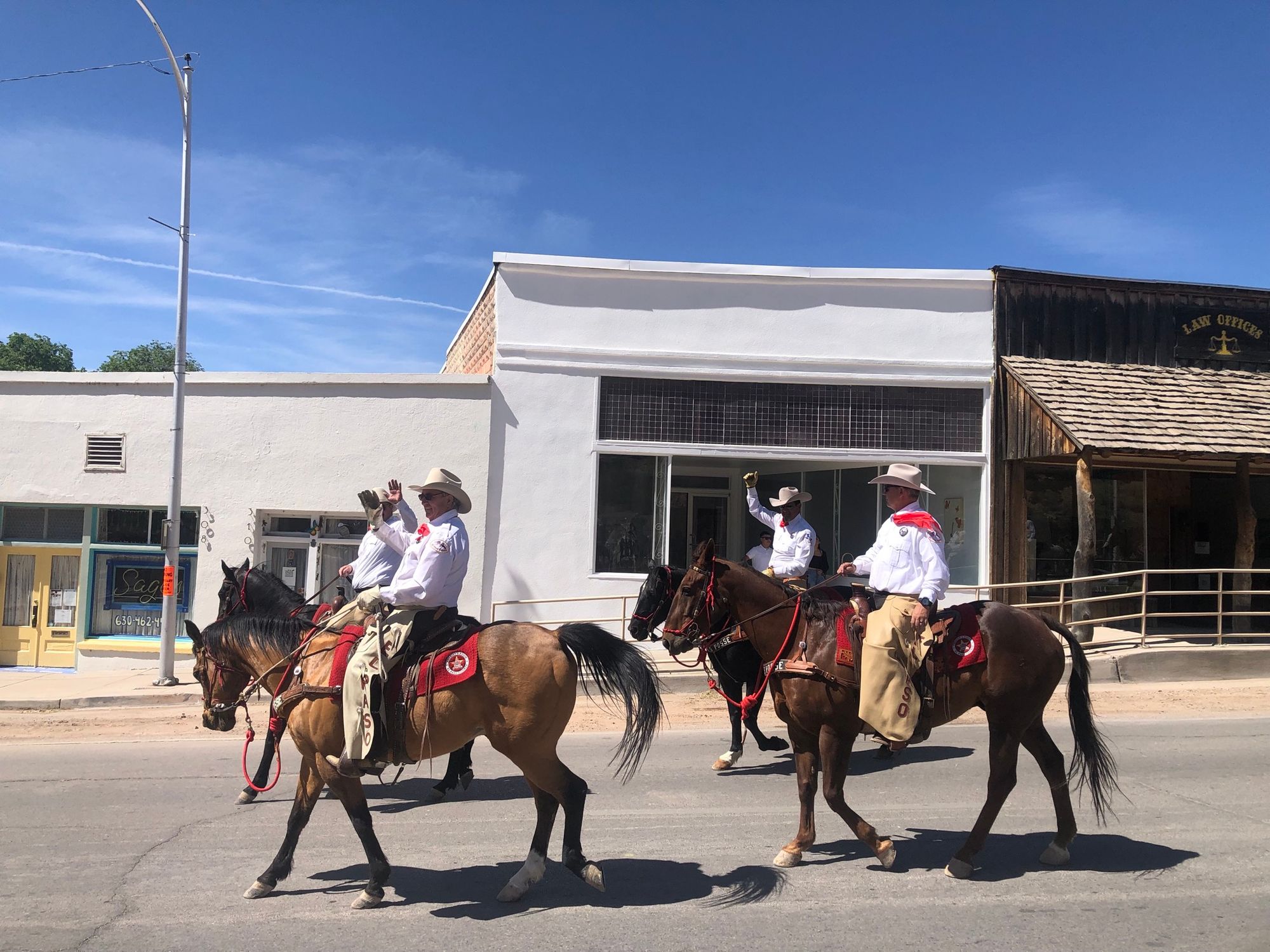 Four men ride horseback, in white shirts, chaps and white cowboy hats, down the Main Street of a small town on a blue sky day