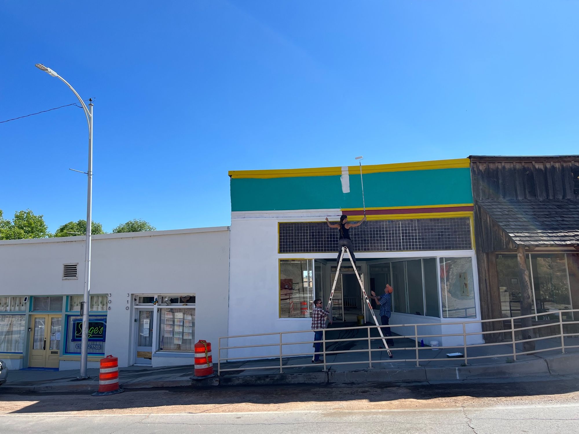 A woman on top of a ladder holds a paint roller and embarks on painting the top of a building.