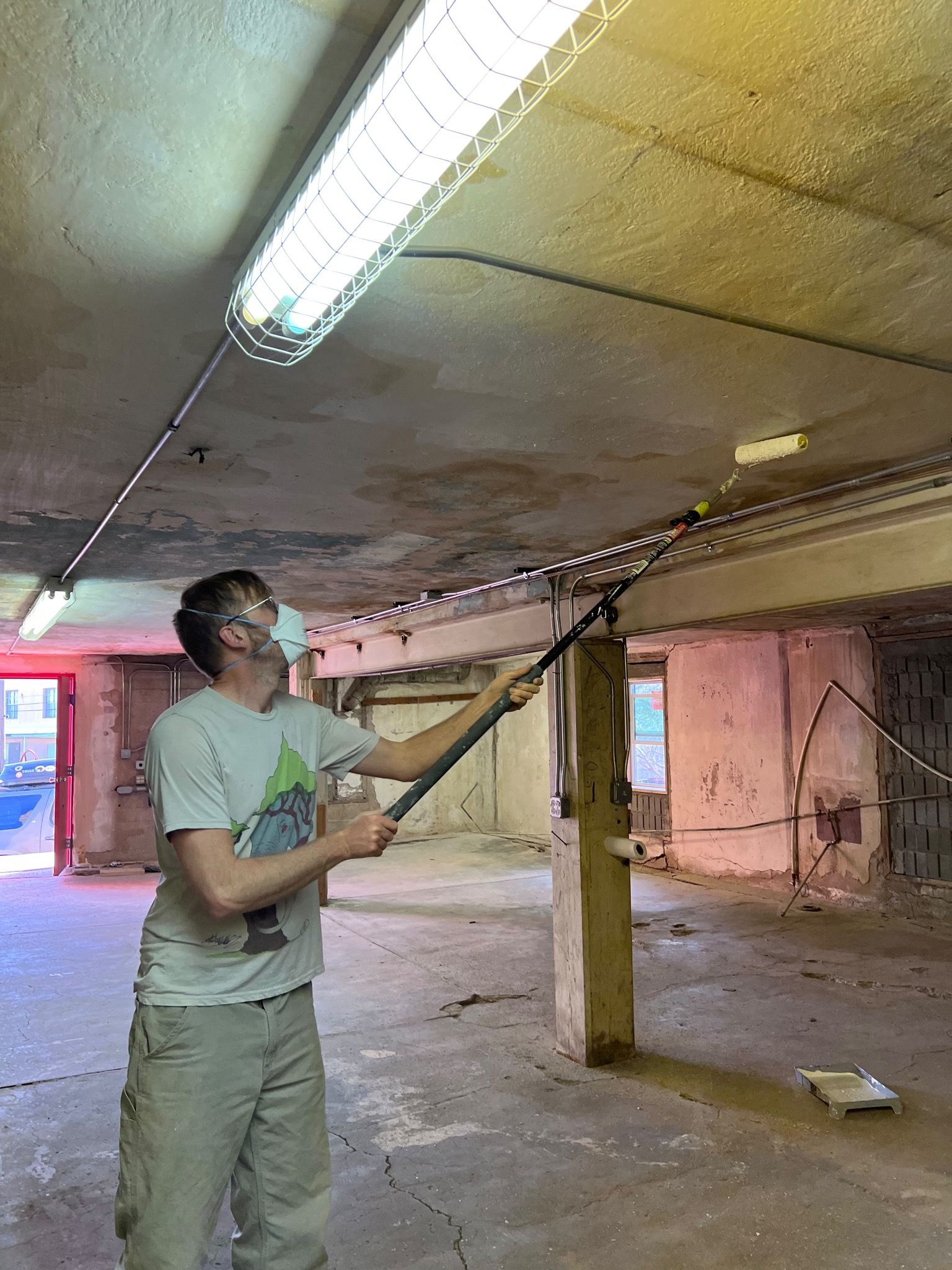 A man stands in an empty building rolling paint on the ceiling. 