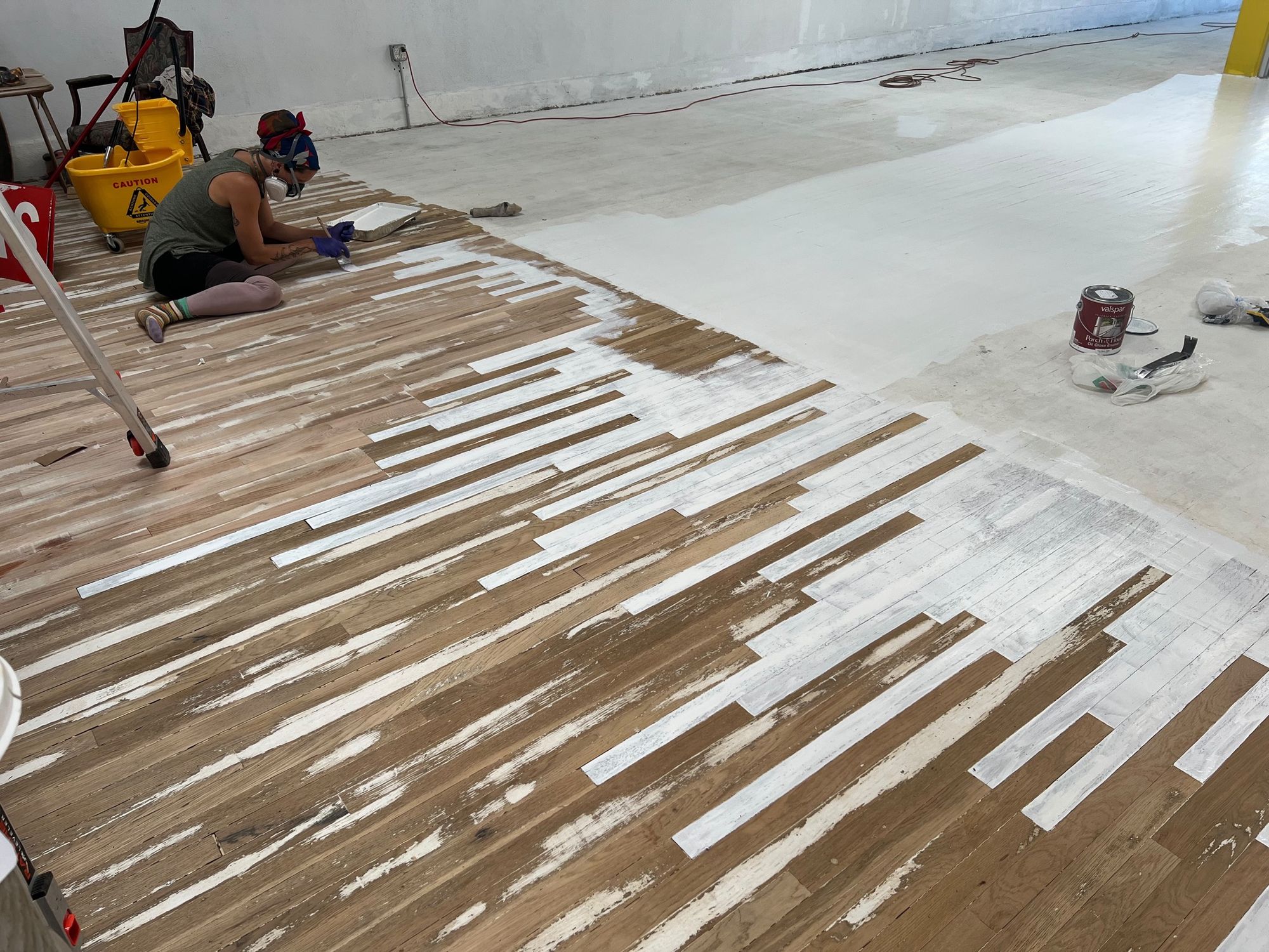 A woman sits on a floor painting individual floor boards white.