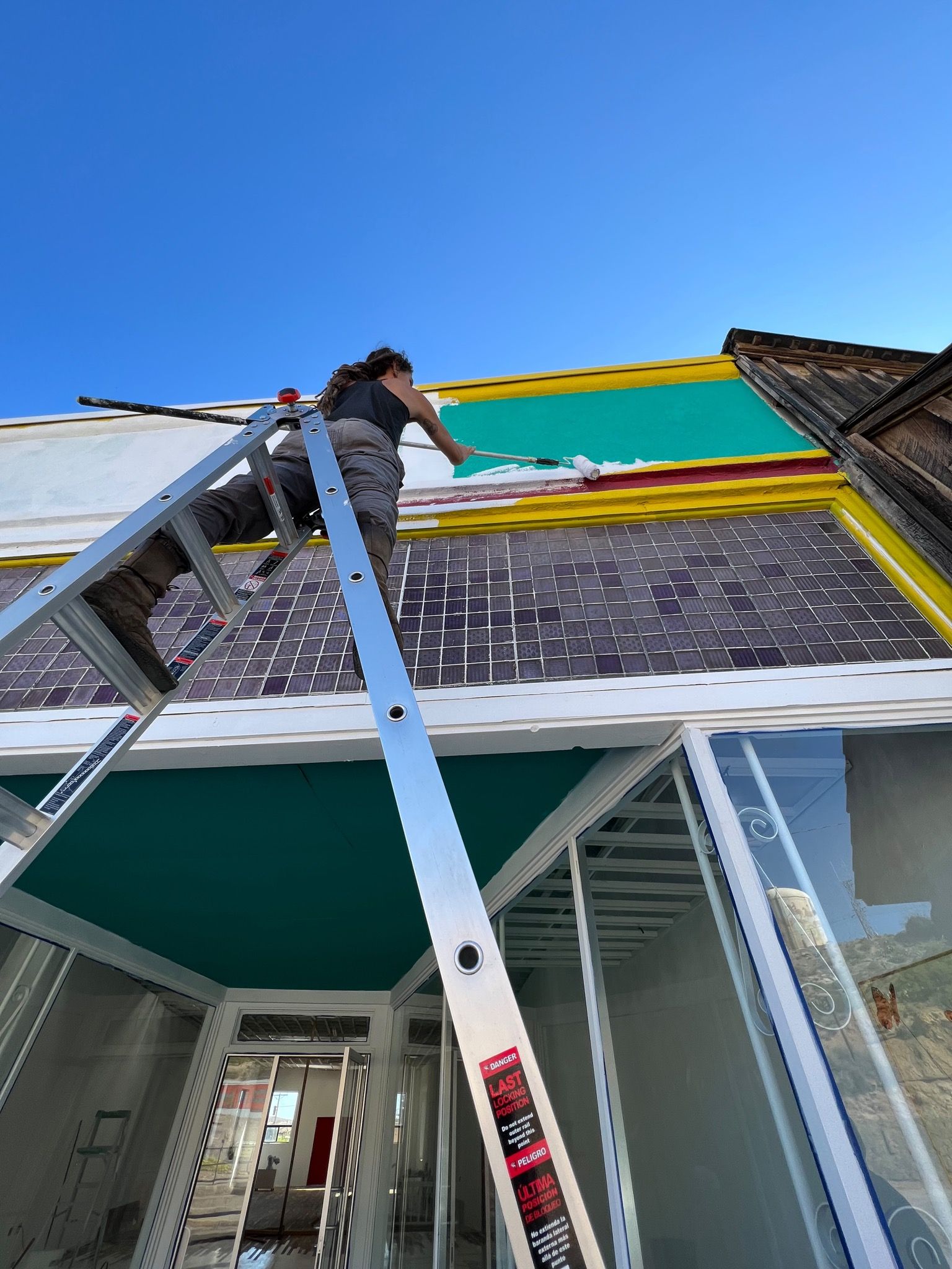 A woman stands on top of a ladder painting the upper edge of a building.