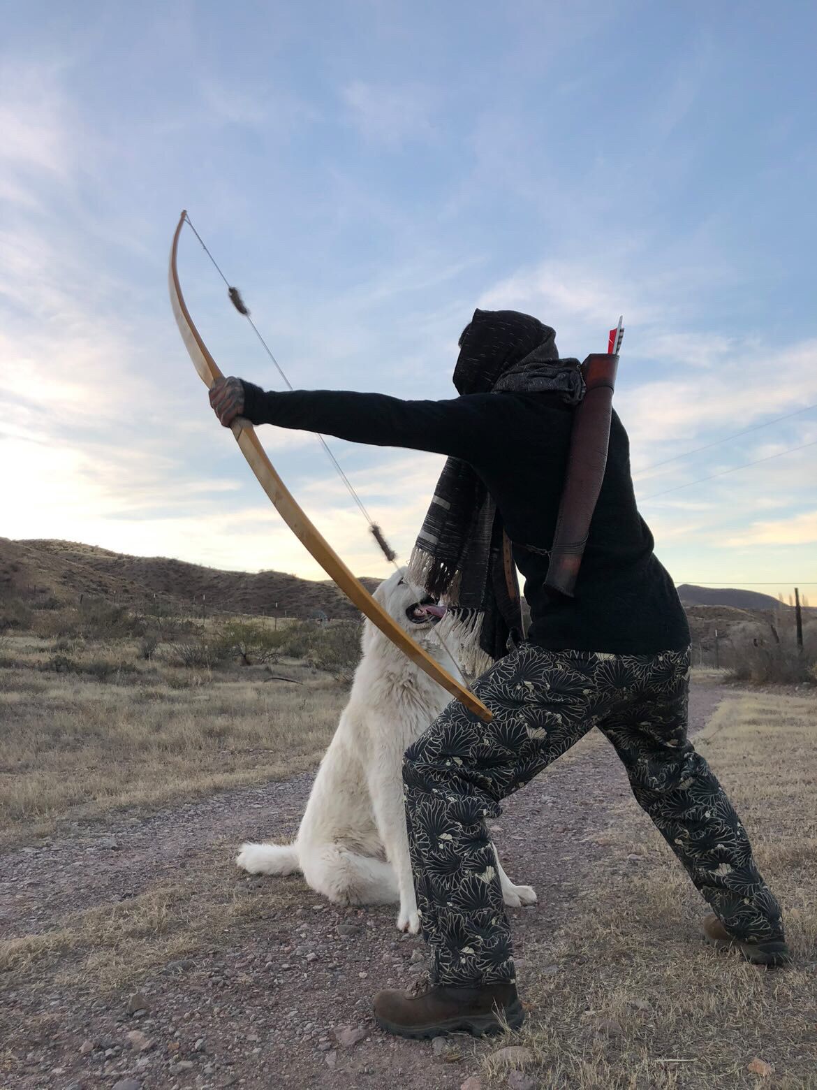 A person wearing all black with a black shawl around their head stands in a warrior stance with a bow and arrow and large fluffy white dog in front of a cactus in the desert